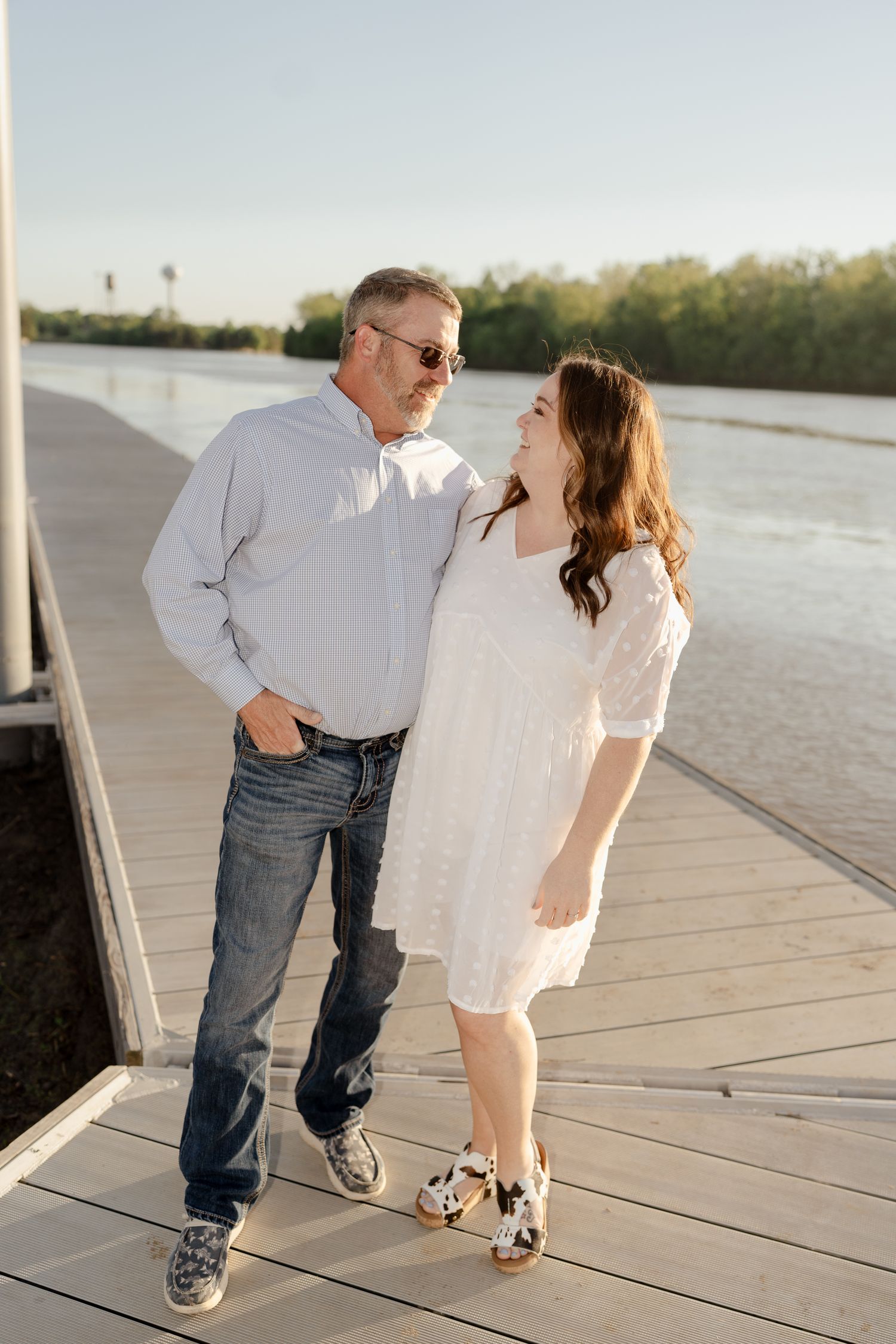 A couple smiles at each other while standing on a wooden dock by the water.