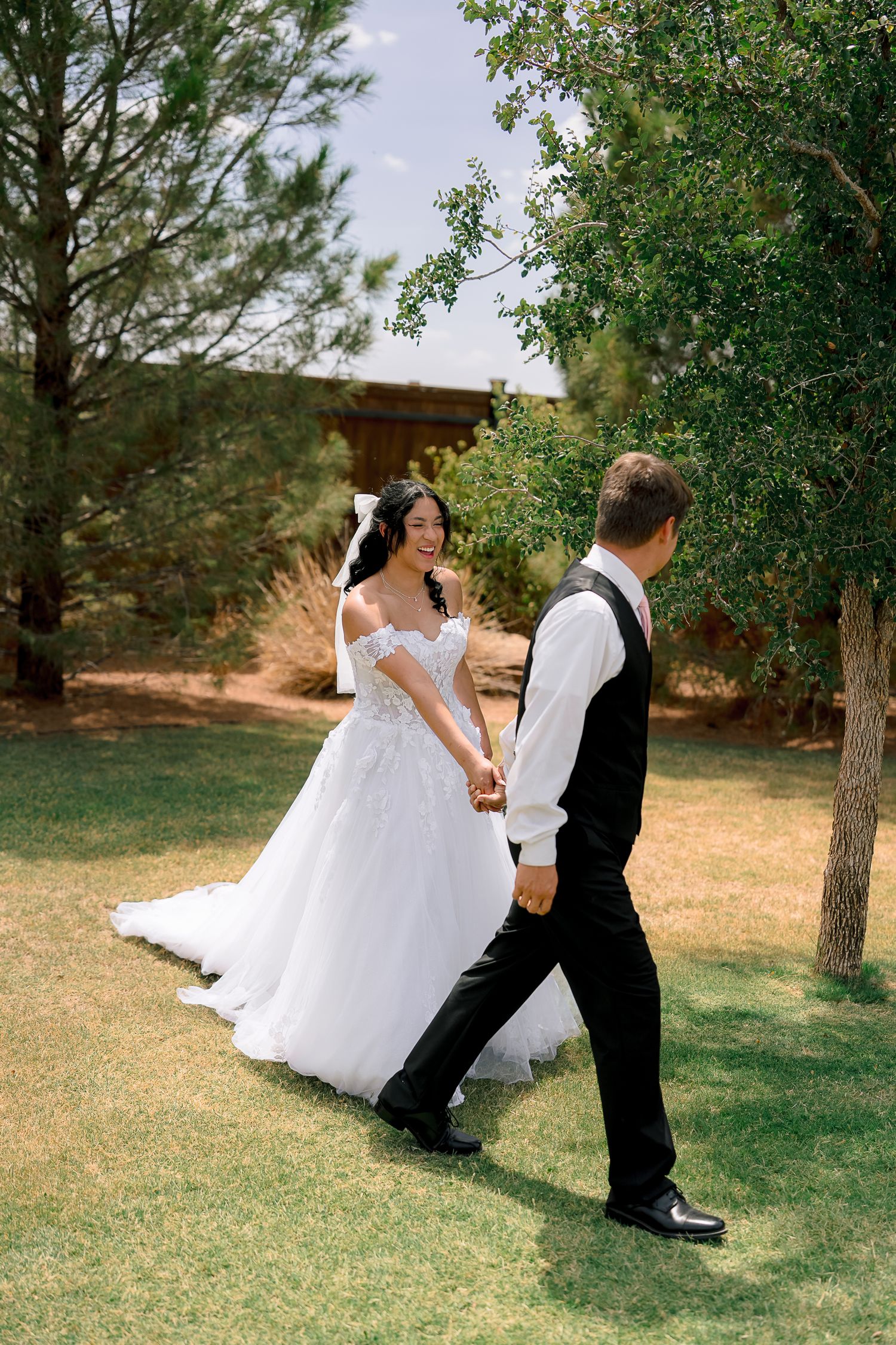 A smiling bride in a white off-shoulder gown holds hands with her groom in a black suit while walking through a garden.