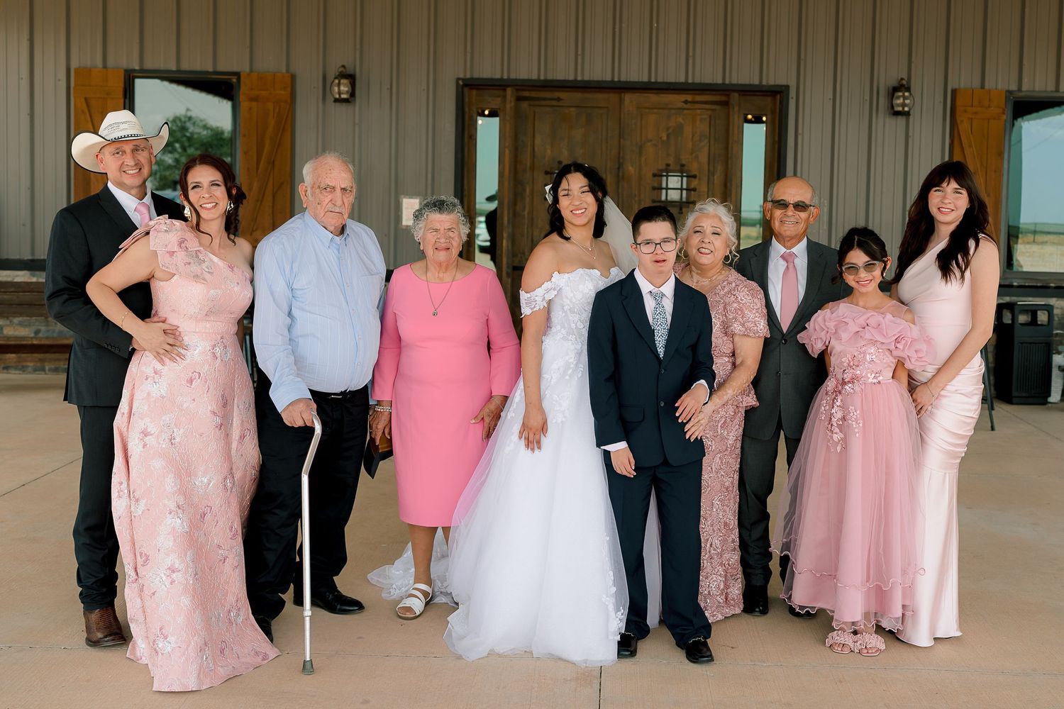 Wedding family portrait with bride in white gown and guests in pink dresses outside rustic barn venue.