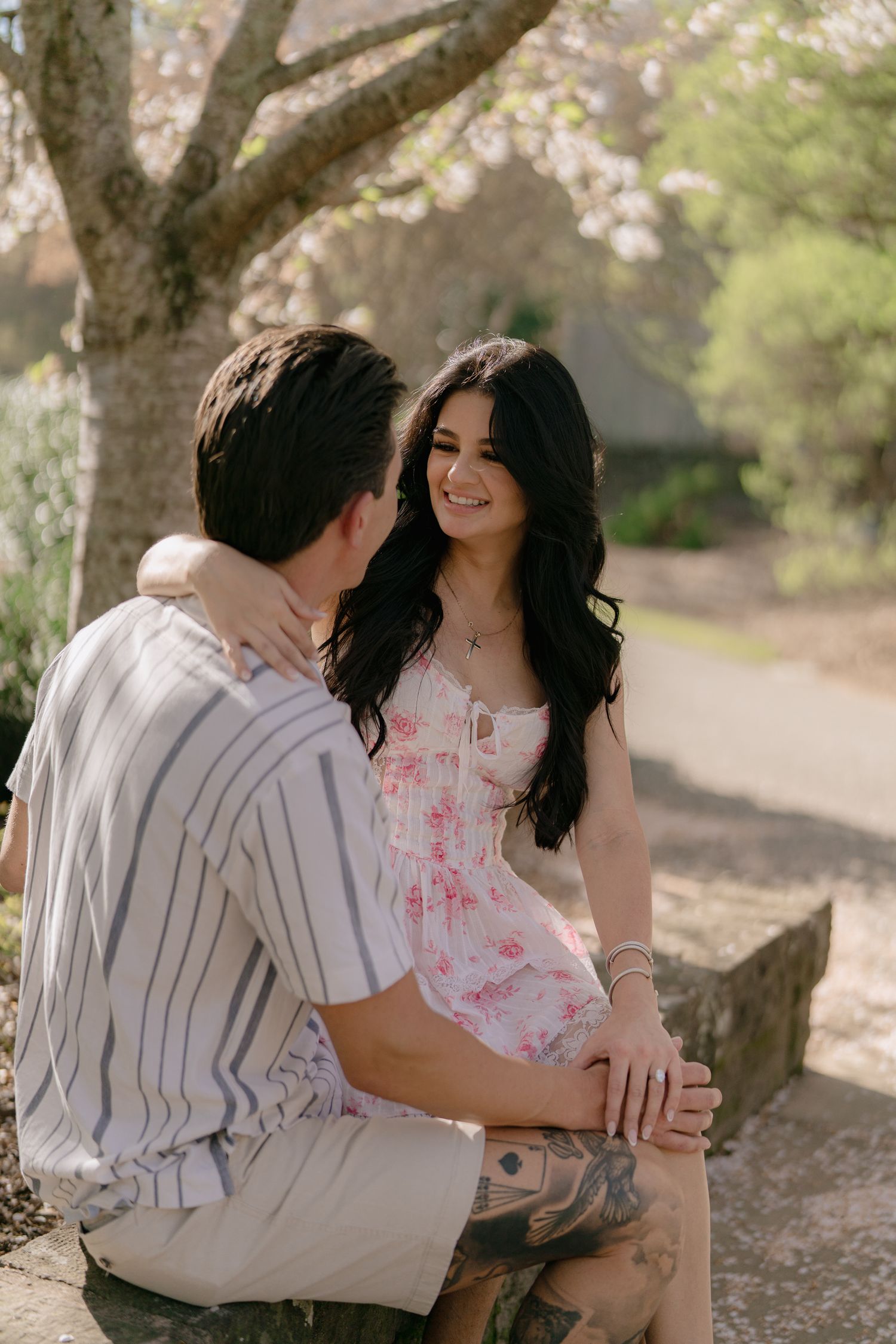 Young woman with long dark hair smiles at tattooed man while sitting on stone bench in spring garden.
