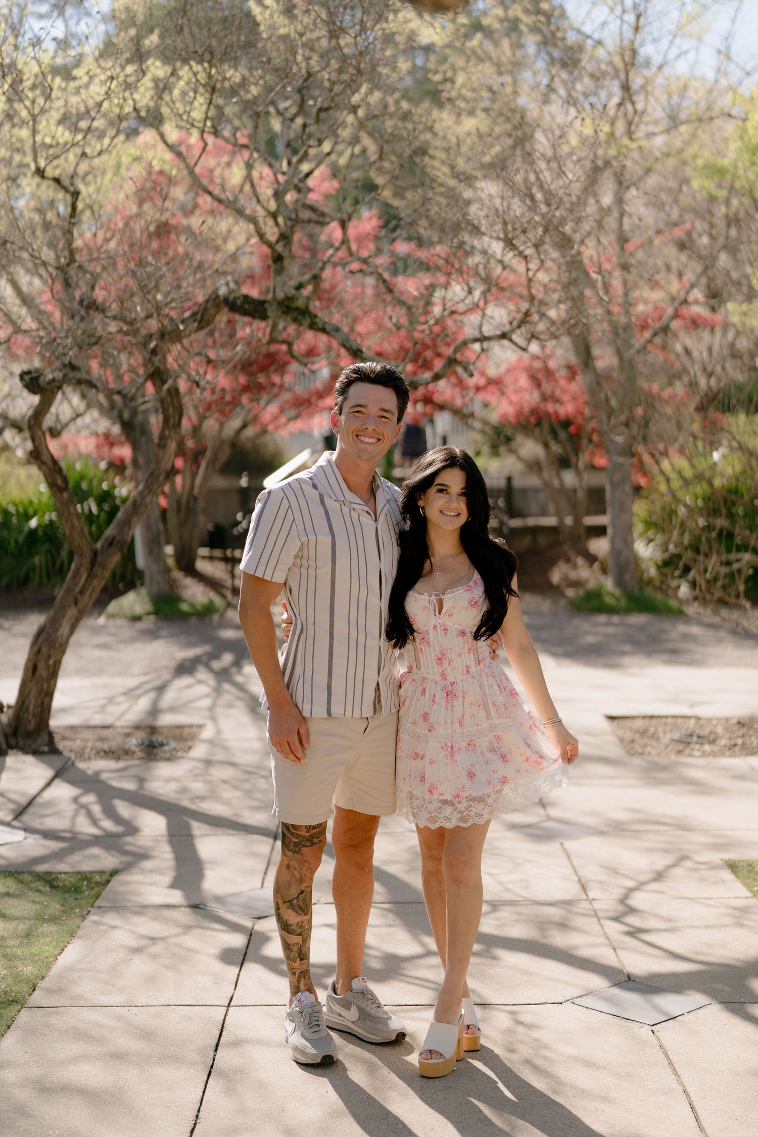 Couple posing on a sunny walkway surrounded by blooming pink trees, man in striped shirt and woman in floral dress.