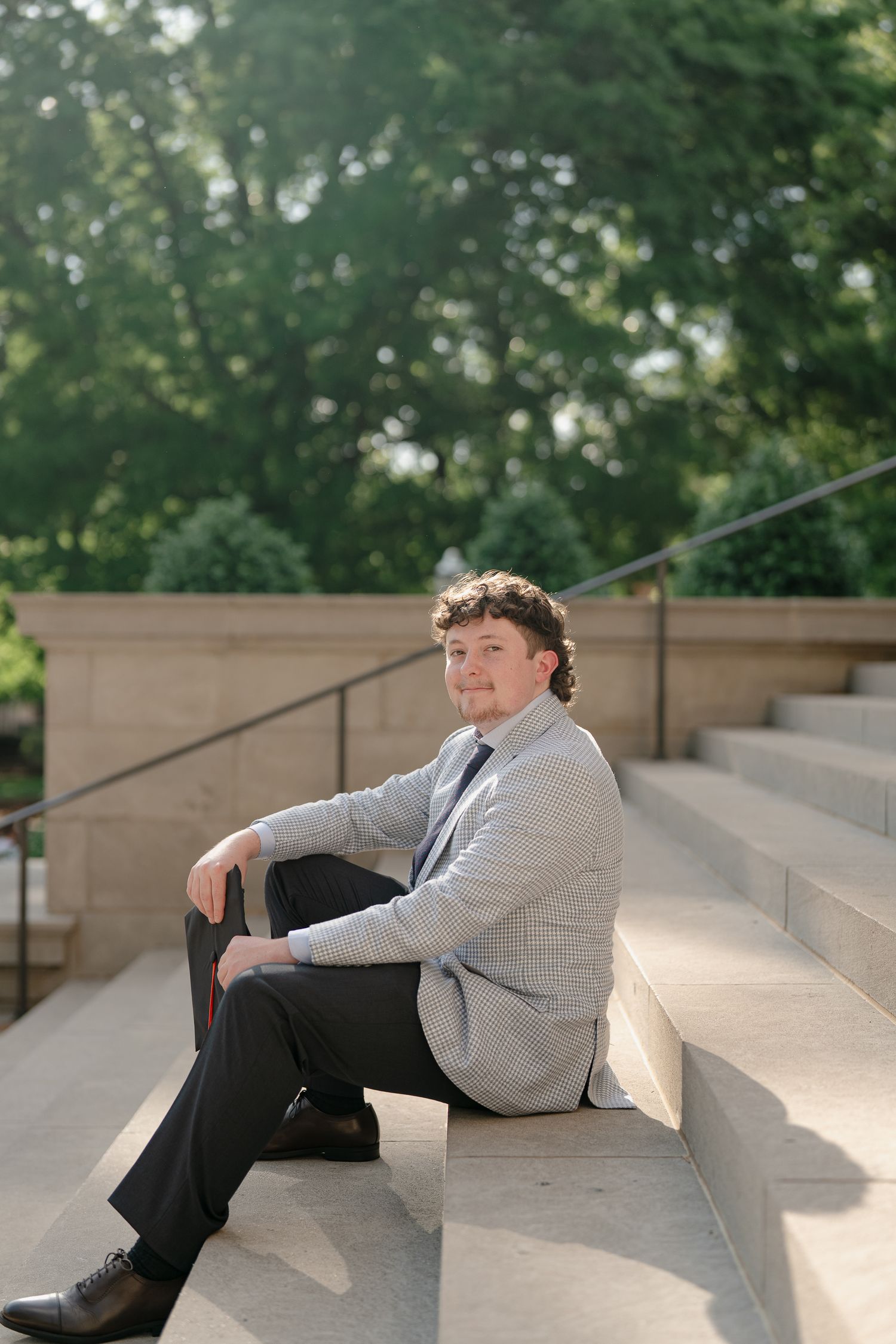 Young man in gray blazer and dark pants sits relaxed on outdoor stone steps surrounded by lush green trees.