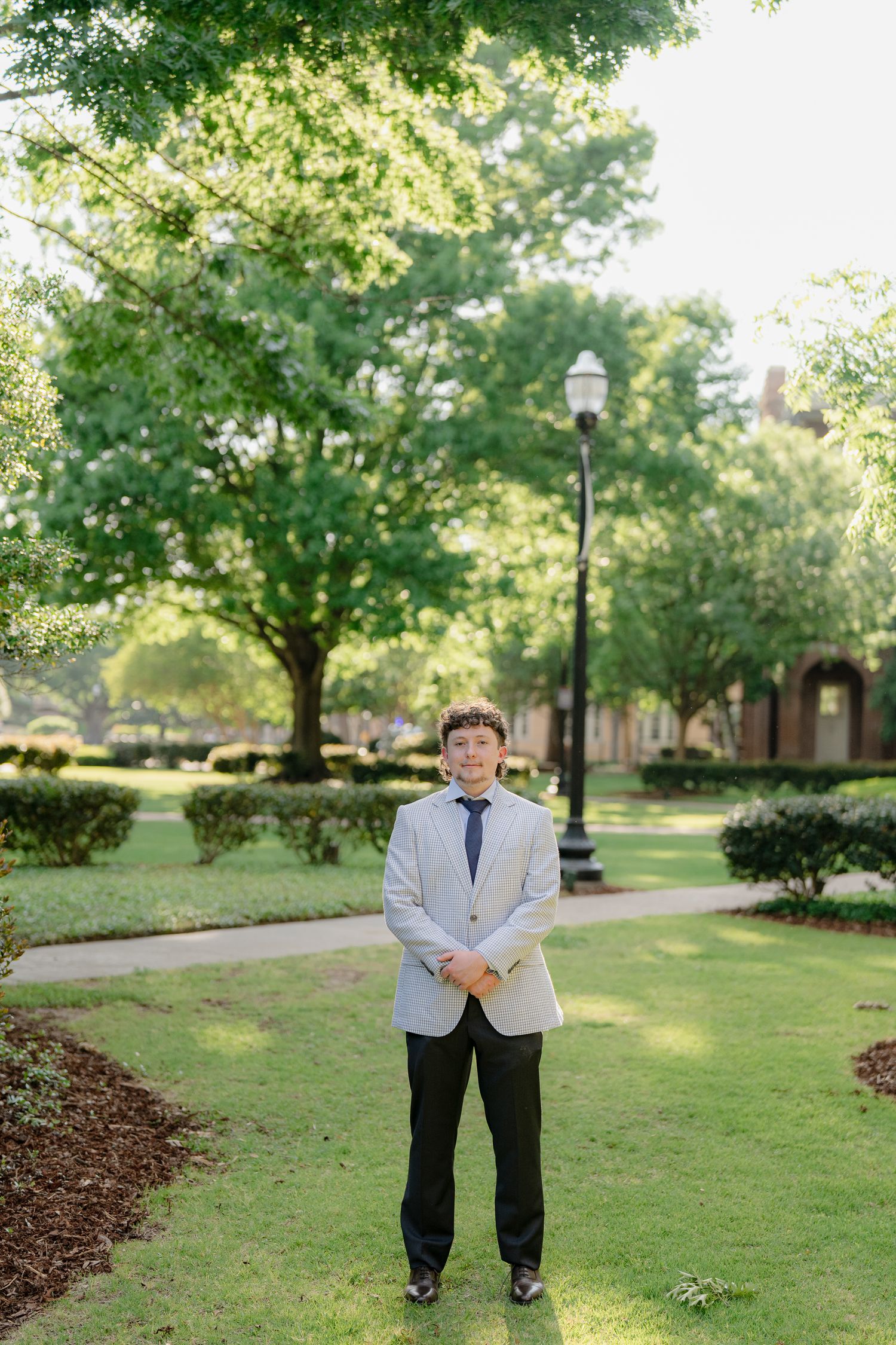 Young man in light blazer and tie stands on a green campus pathway surrounded by lush trees.