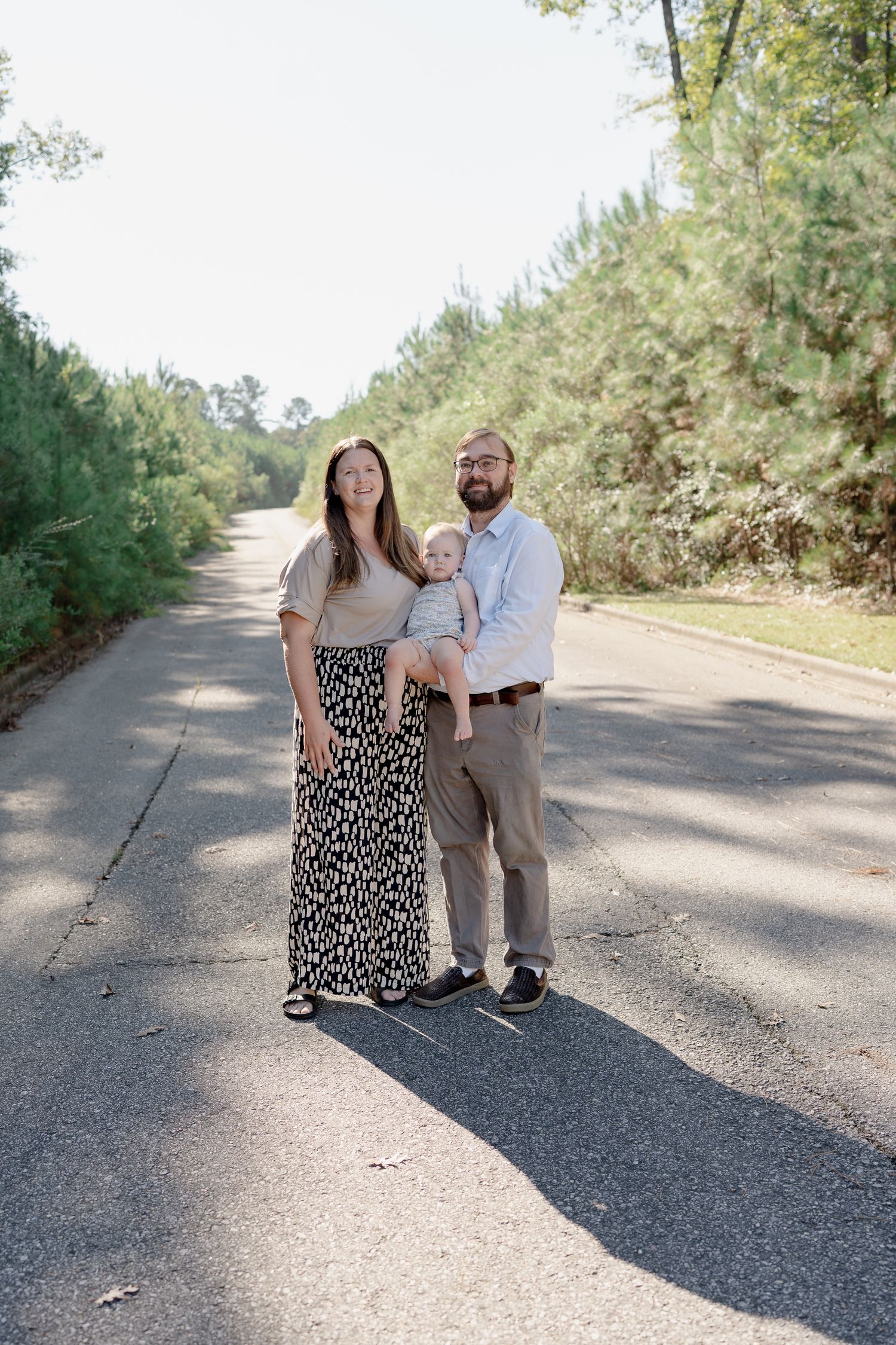 A couple stands together on a tree-lined gravel path on a sunny day, wearing coordinated casual attire.