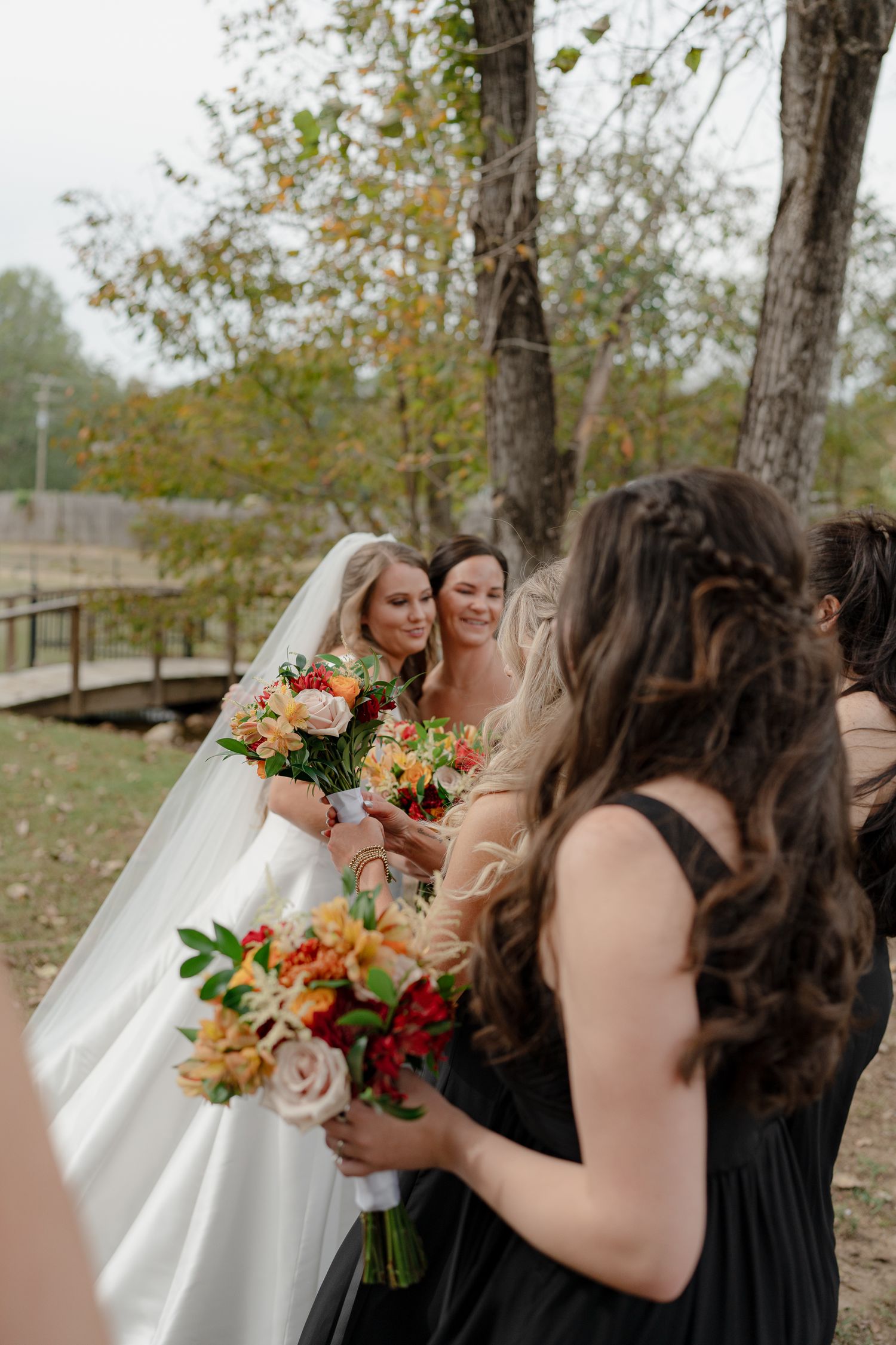 Bridesmaids in black dresses gather around bride holding colorful fall bouquets outdoors.