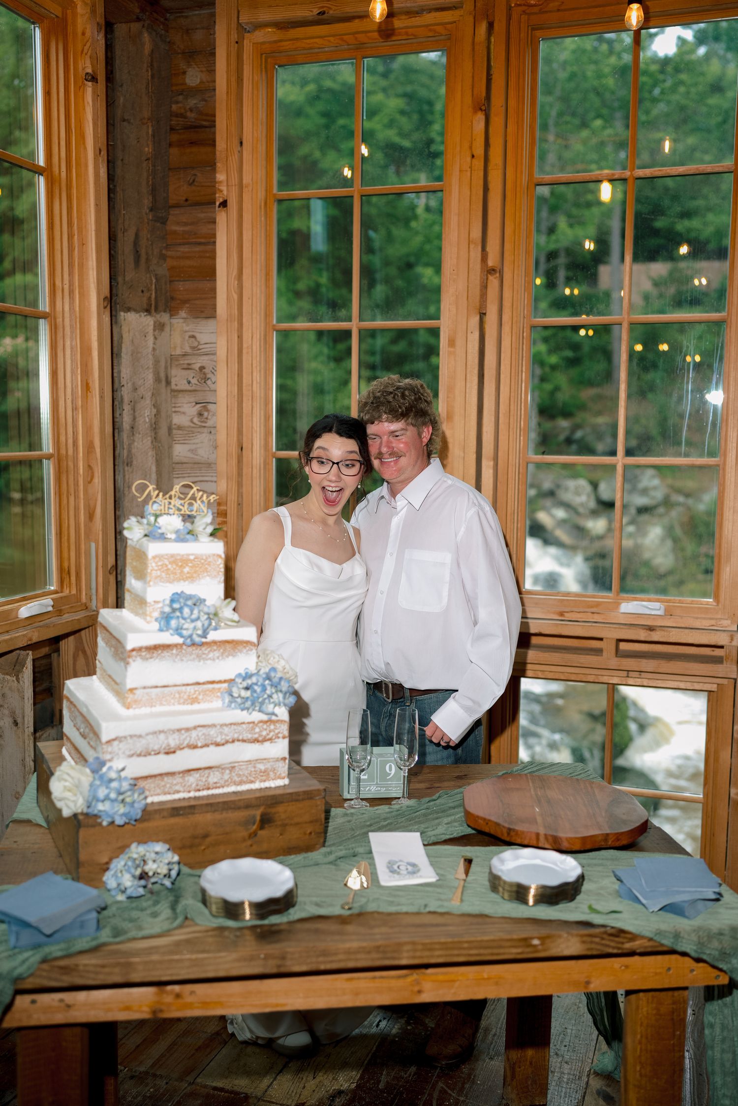 Bride and groom smiling beside a tiered wedding cake decorated with blue hydrangeas in a rustic wooden venue.