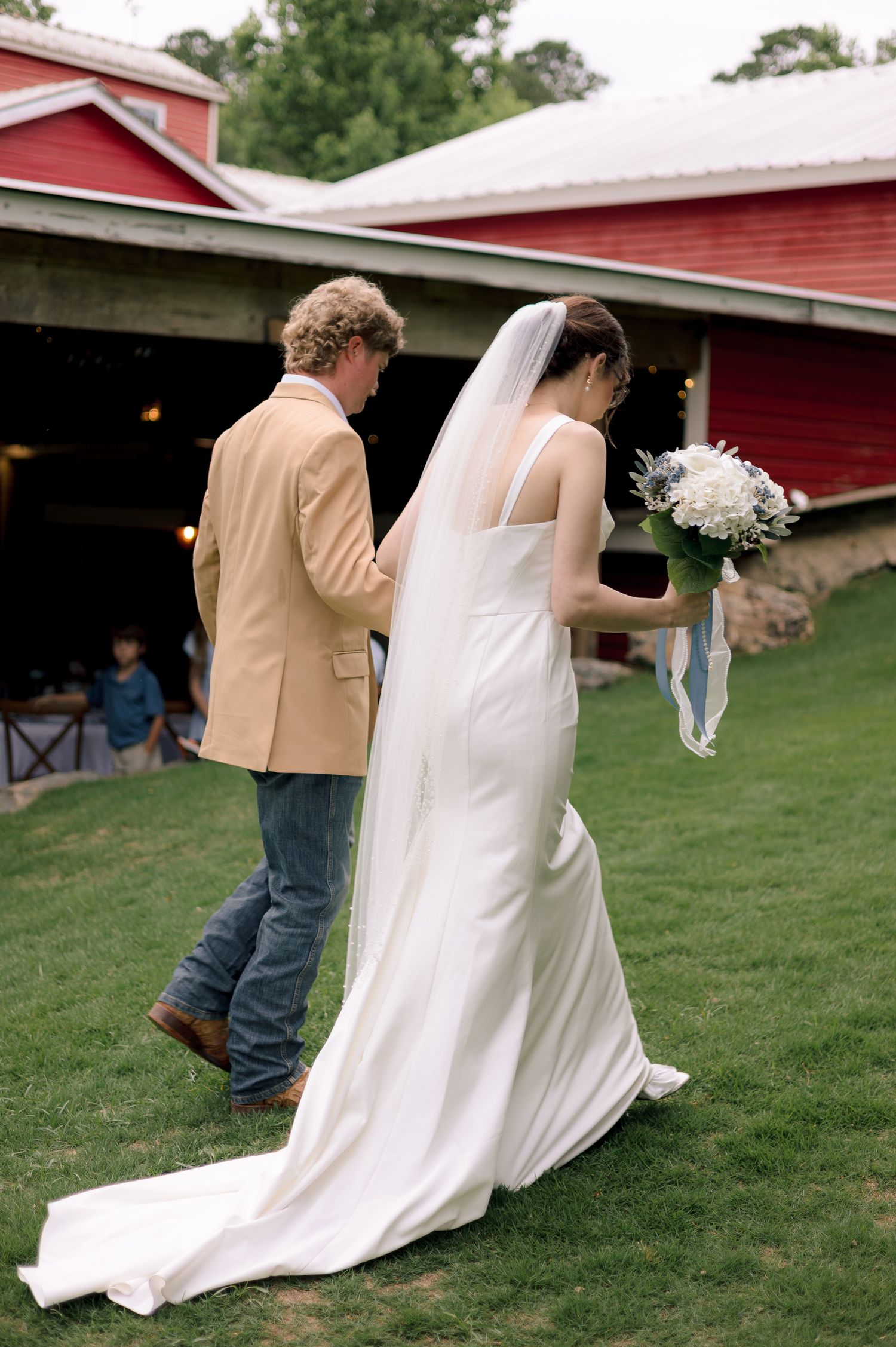 Bride in white gown with veil and groom in tan blazer walk toward red barn wedding venue.