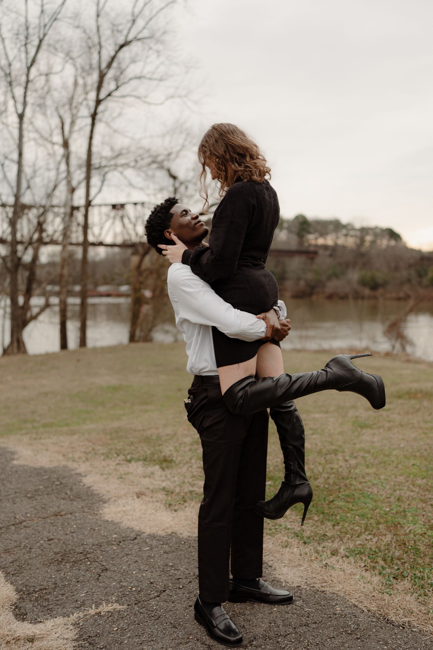 A man lifts his partner into the air near a river, both dressed in black and white, sharing a romantic moment.