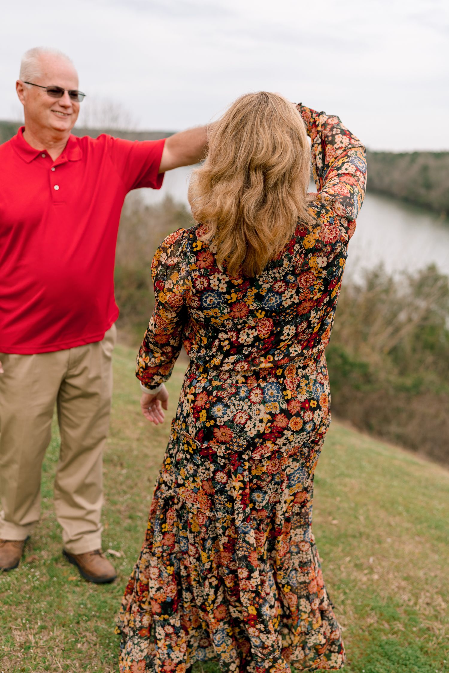 A man in a red polo adjusts a woman's hair outdoors near a scenic river overlook.