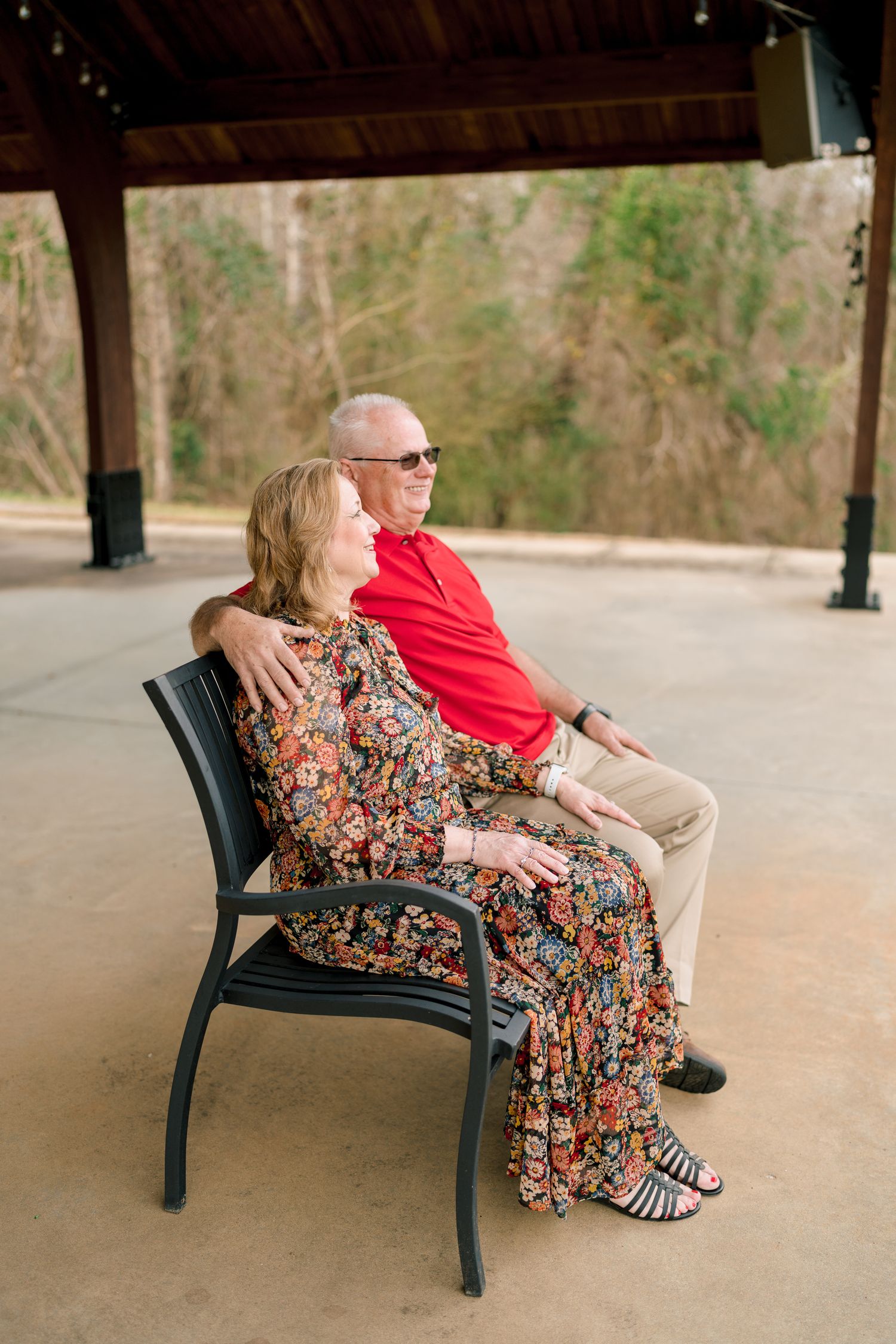 Happy older couple sitting together on bench under pavilion, woman in floral dress and man in red shirt.