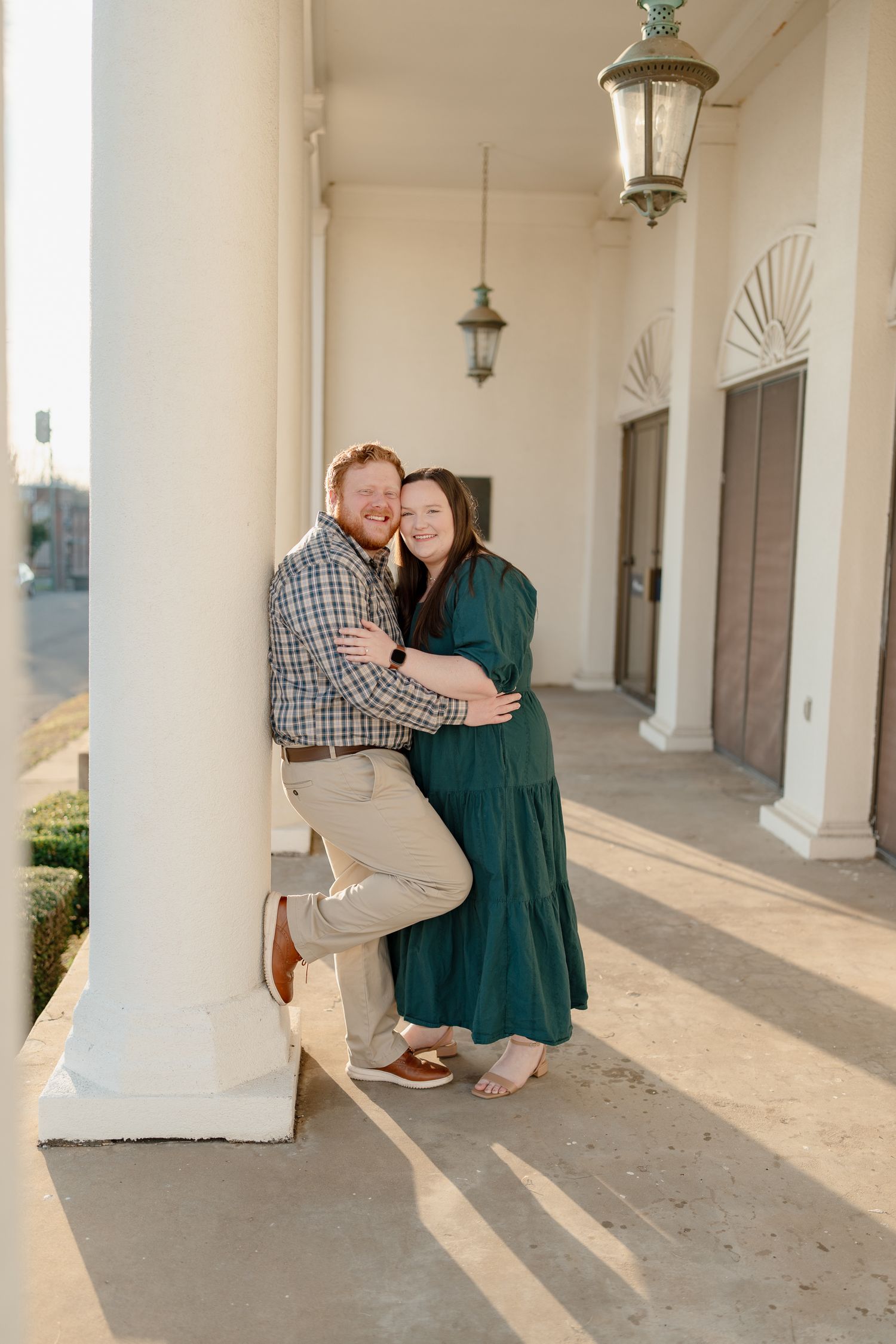 Couple embracing by white column on sunlit porch, woman in green dress and man in plaid shirt smiling together.