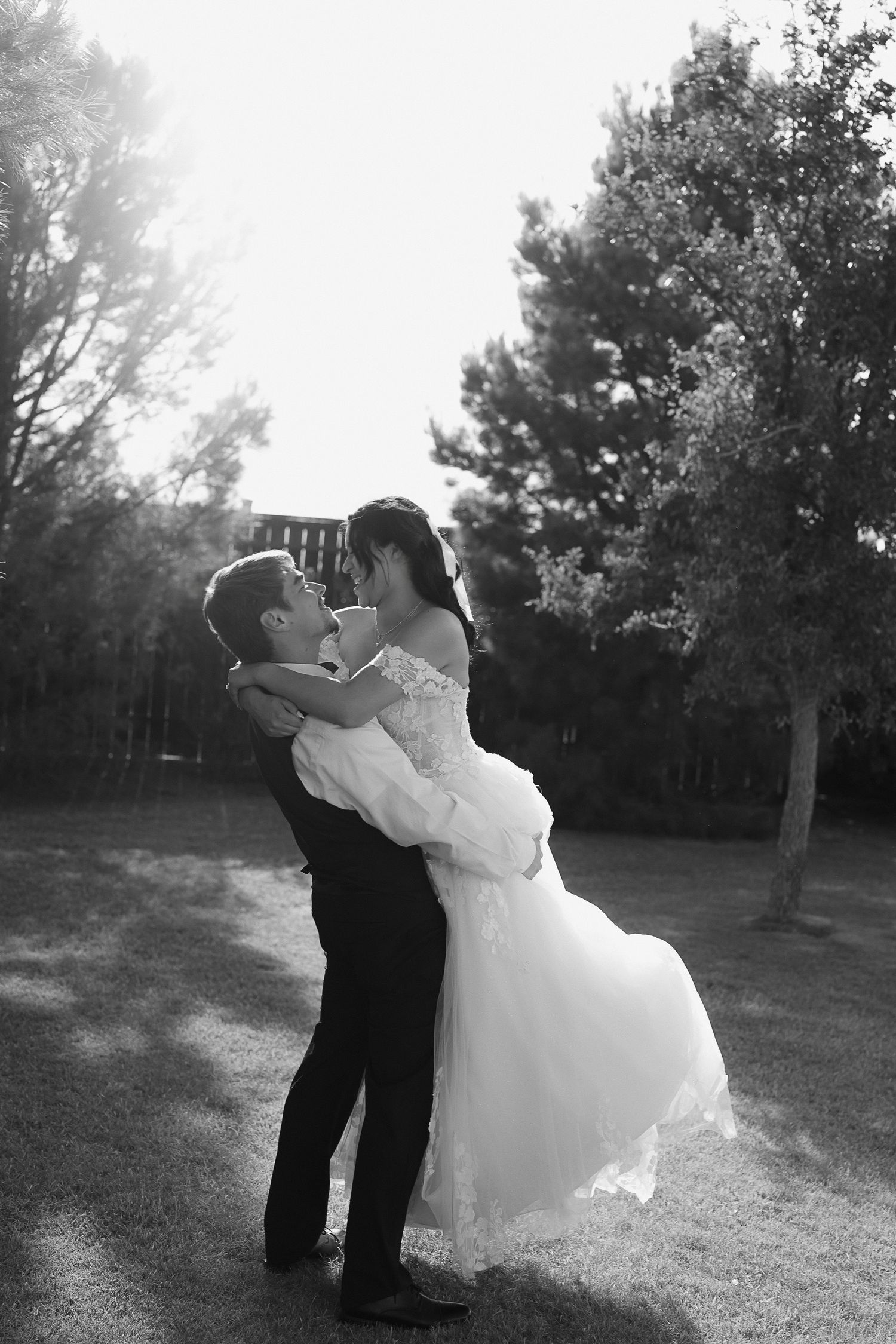 Black and white photo of groom lifting bride in a wedding dress outdoors, sharing a joyful moment among trees.