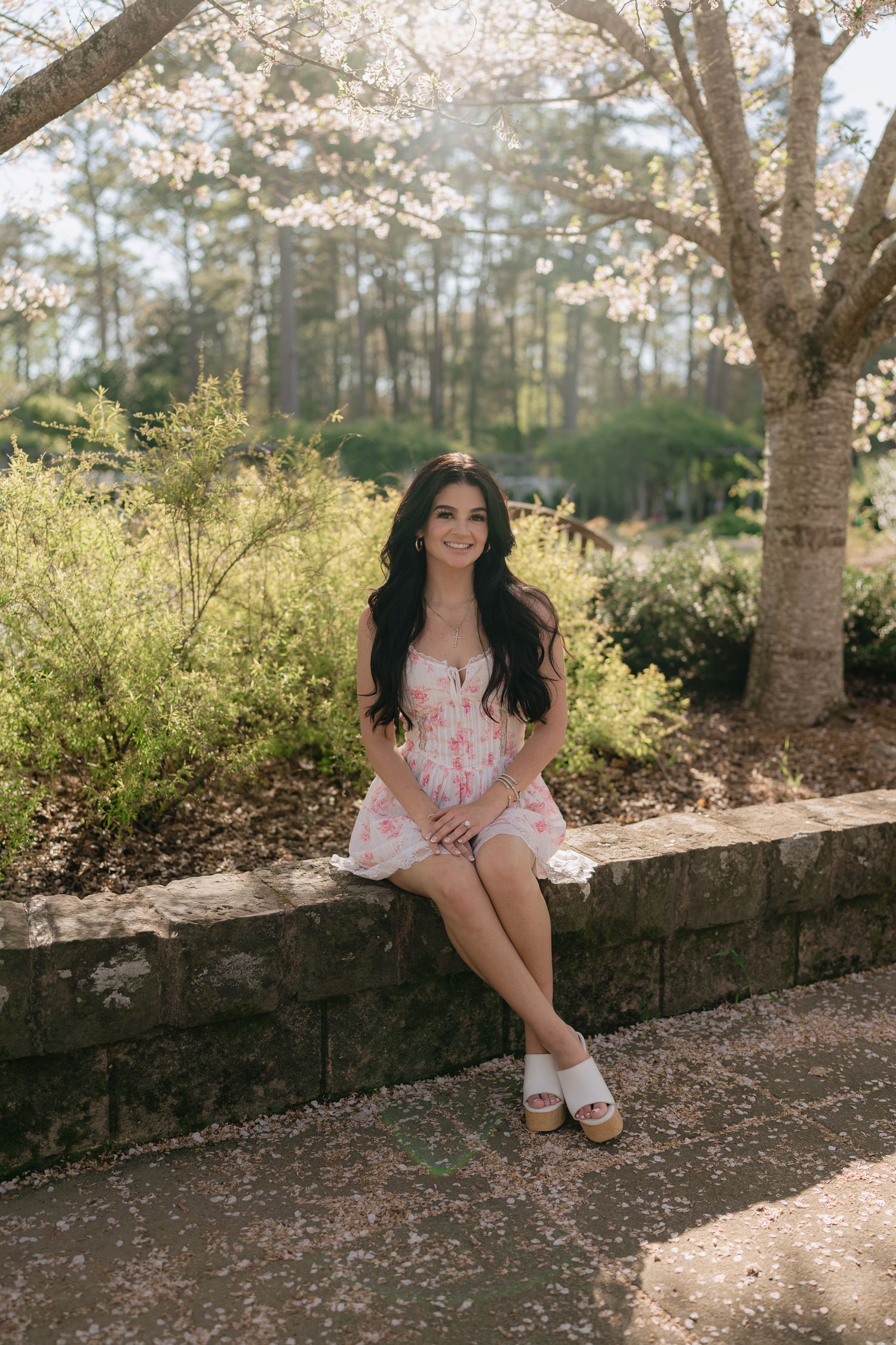 Young woman in a floral pink dress sitting on a stone wall beneath blooming white spring trees.