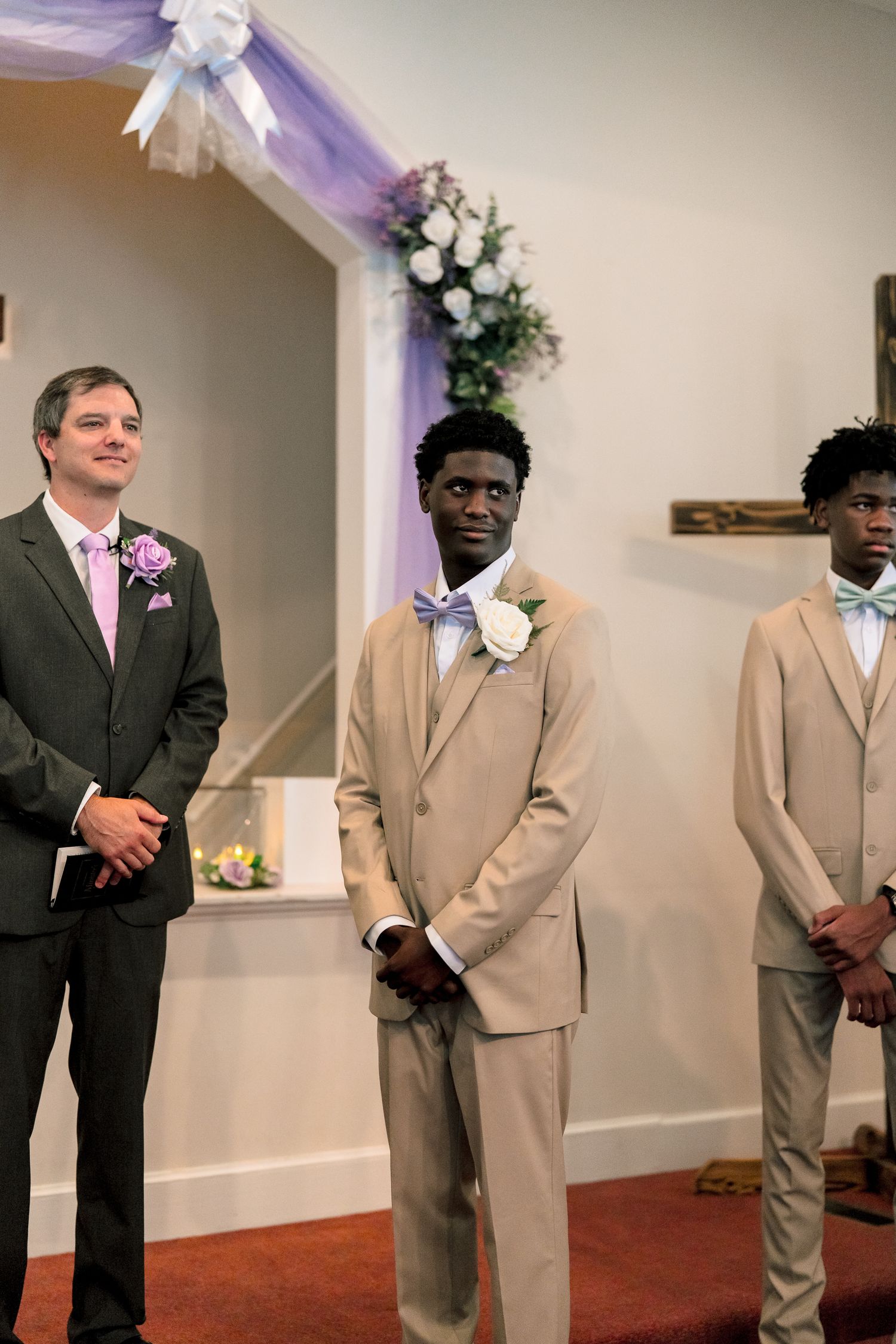 Three groomsmen in tan and charcoal suits with lavender accessories standing at wedding altar in decorated church.