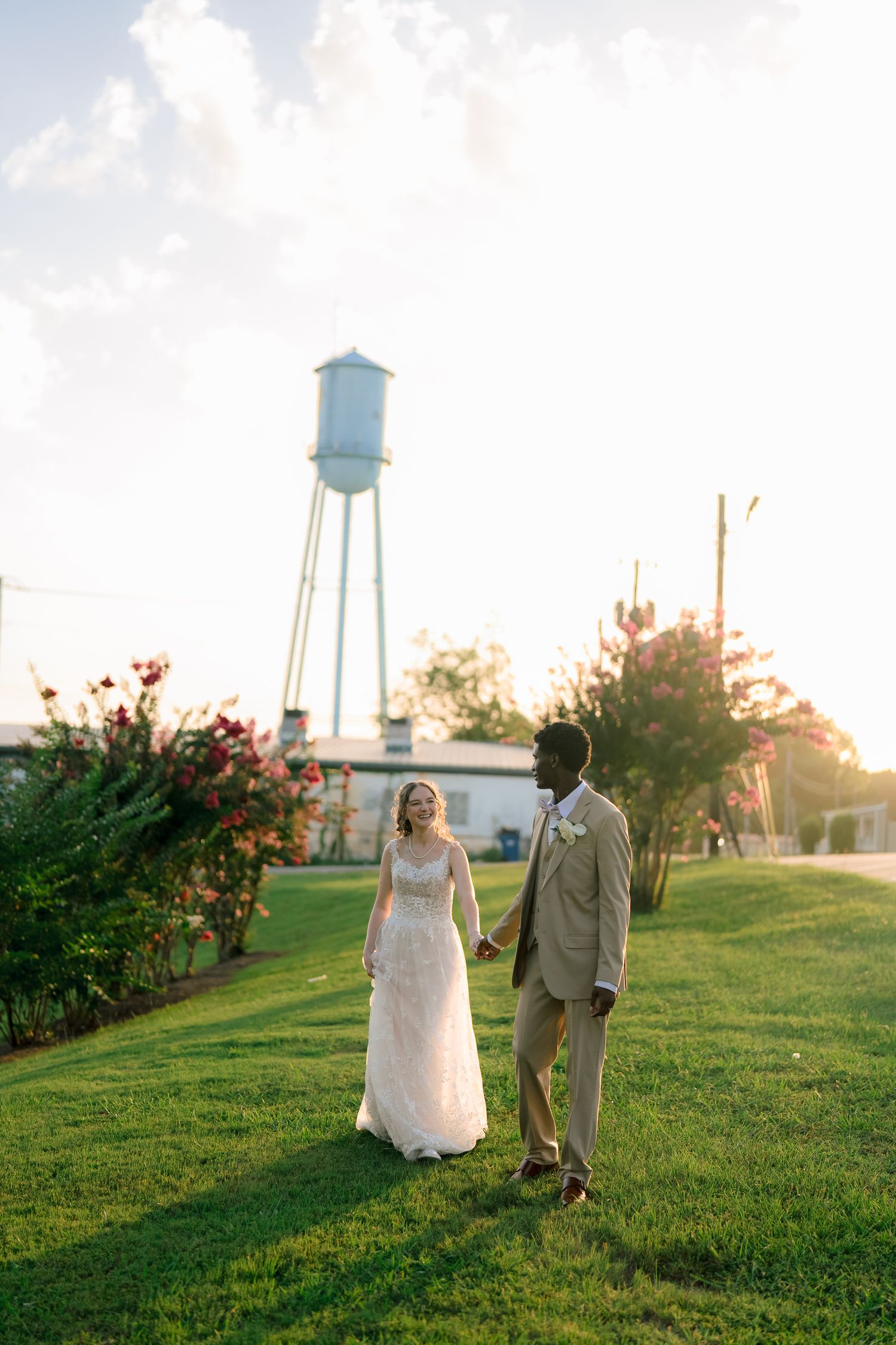 Bride and groom holding hands walking on green lawn with water tower at golden hour.