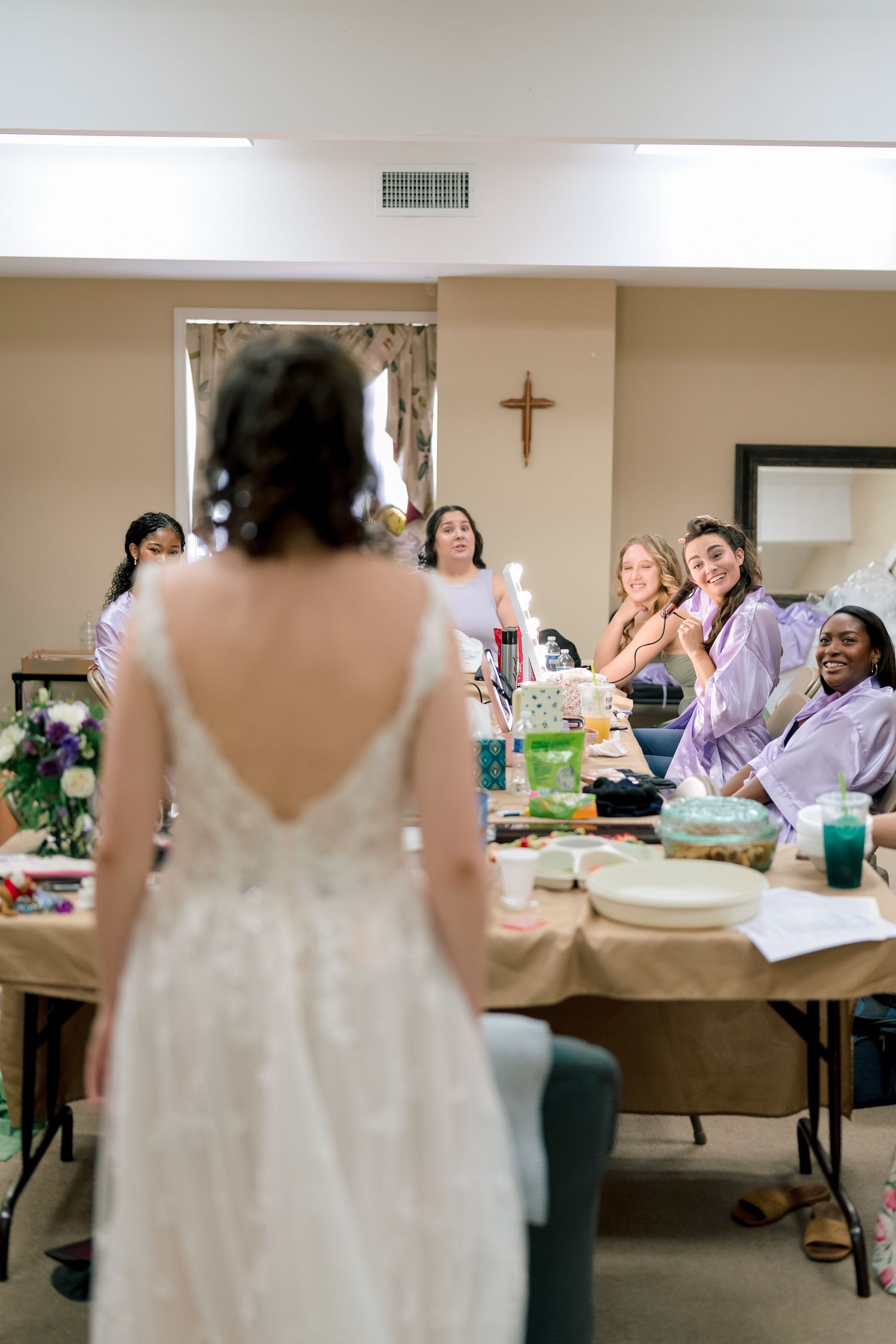 Bride in backless lace wedding gown reveals dress to excited bridesmaids in purple robes seated at table.