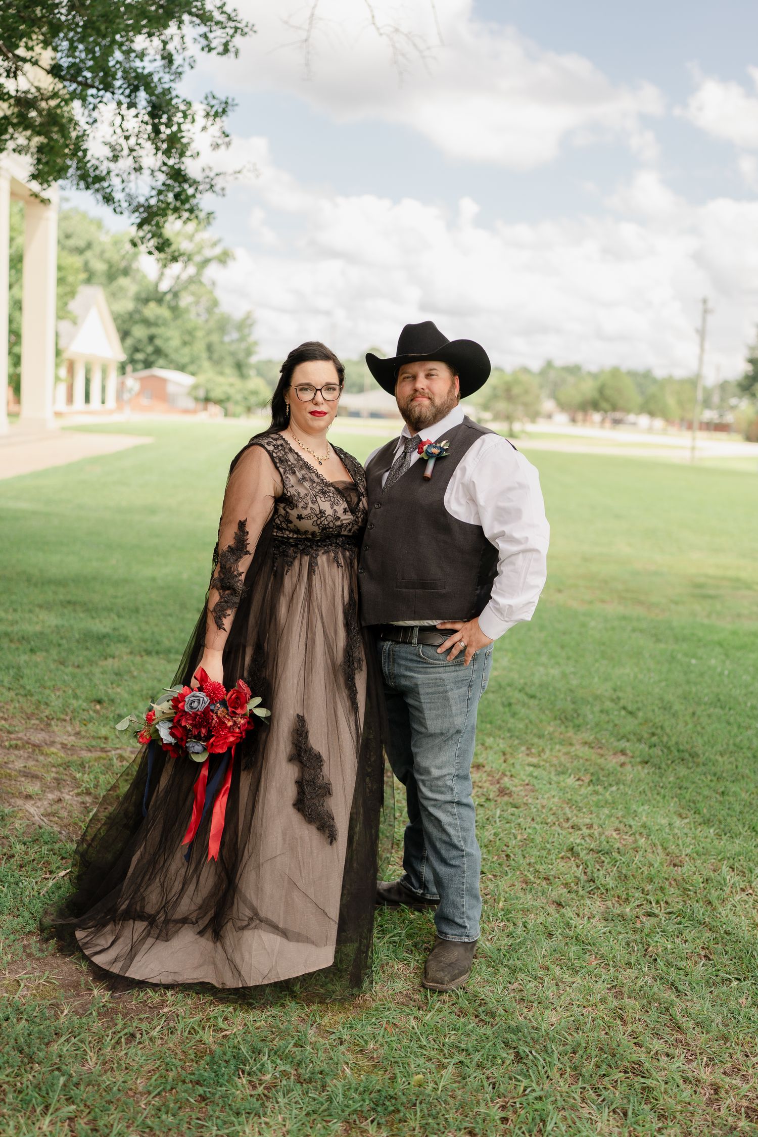 A couple poses together on grassy grounds wearing western and formal attire.