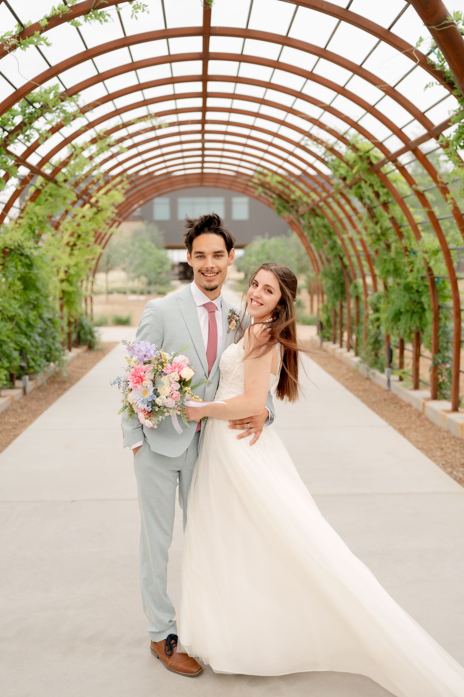 Newlyweds share joyful moments in a rustic greenhouse tunnel with arched wooden beams.