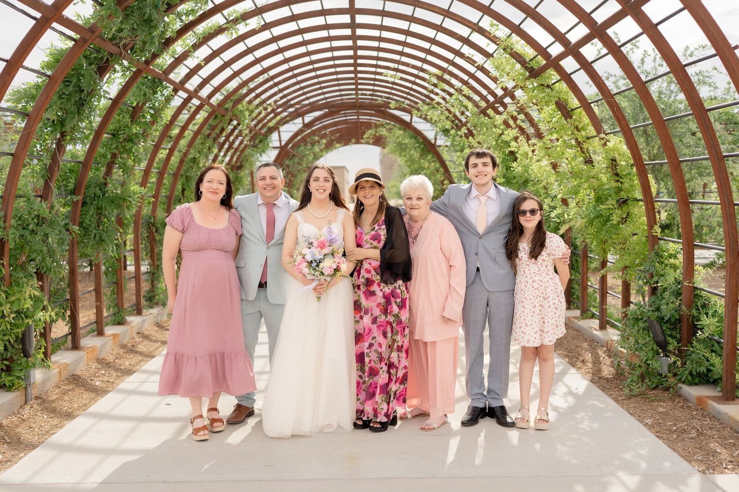 Wedding party poses in an elegant wooden arched garden tunnel with lush greenery overhead.