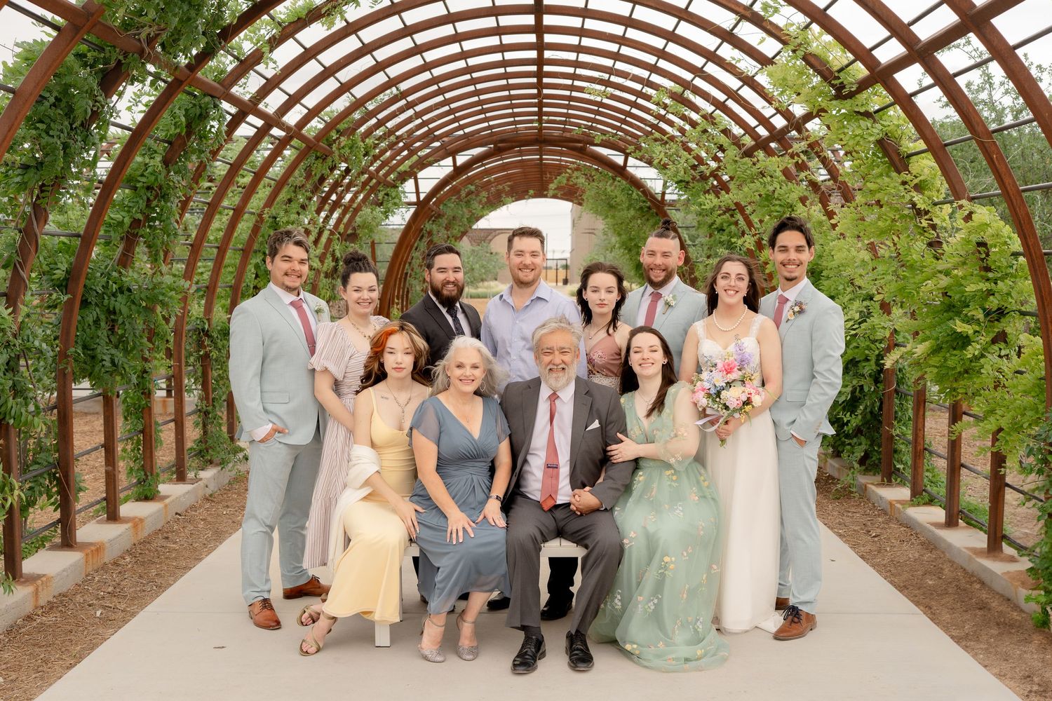 Wedding party poses together under a wooden arched garden tunnel covered in greenery and natural light.