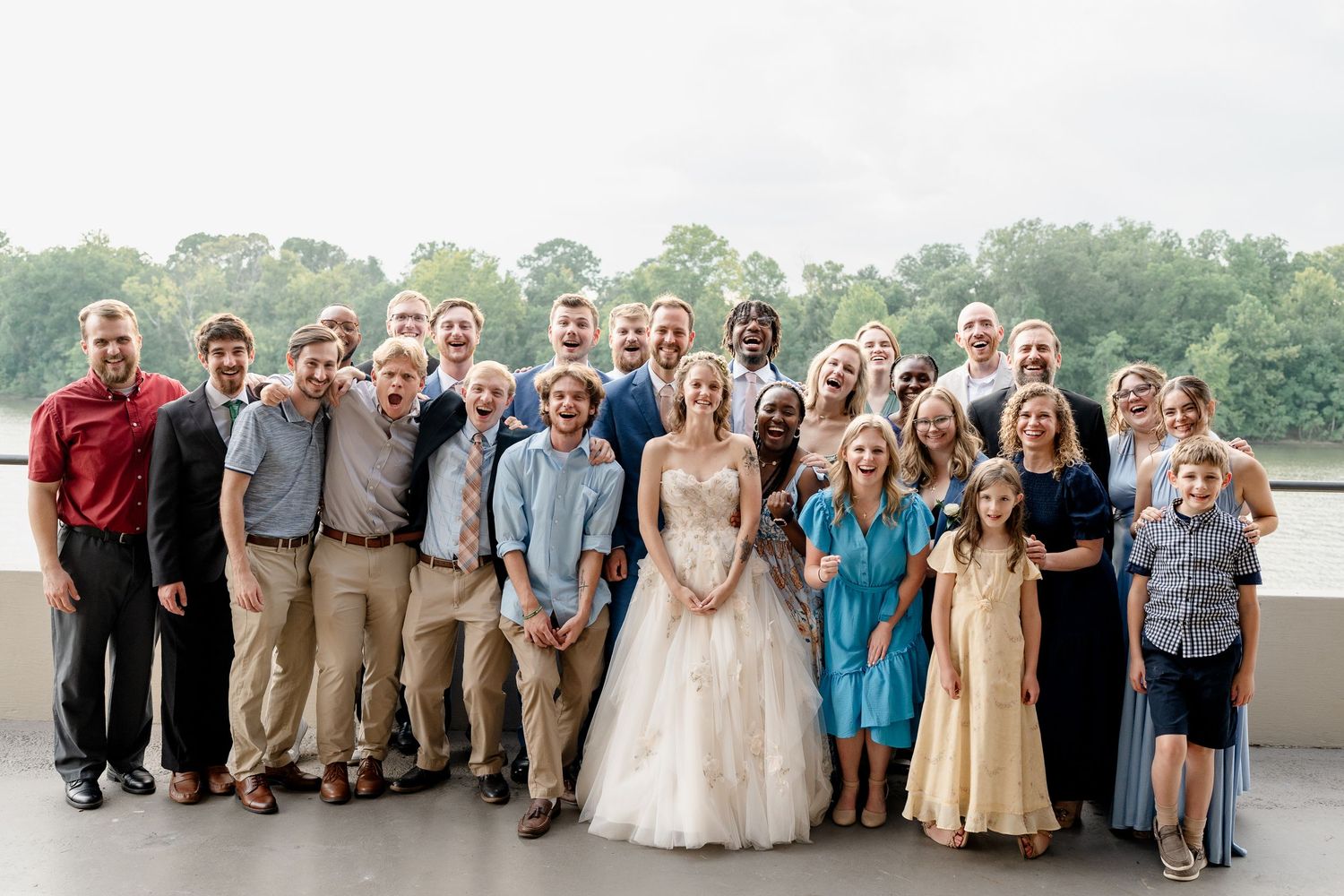 A wedding party group poses together outdoors near a waterfront on a cloudy day.