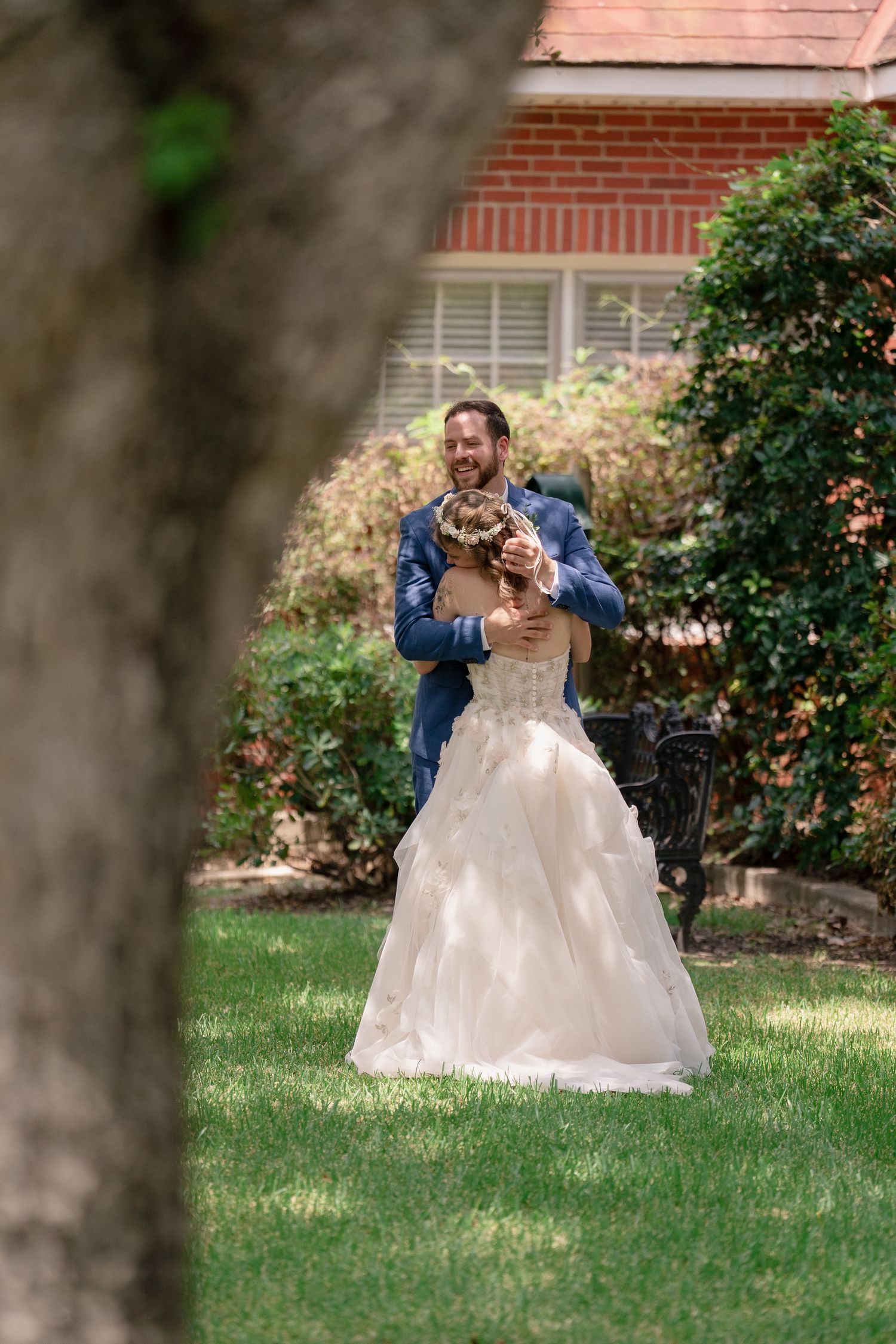 A romantic wedding scene unfolds on a grassy lawn with flowering bushes and brick architecture in the background.
