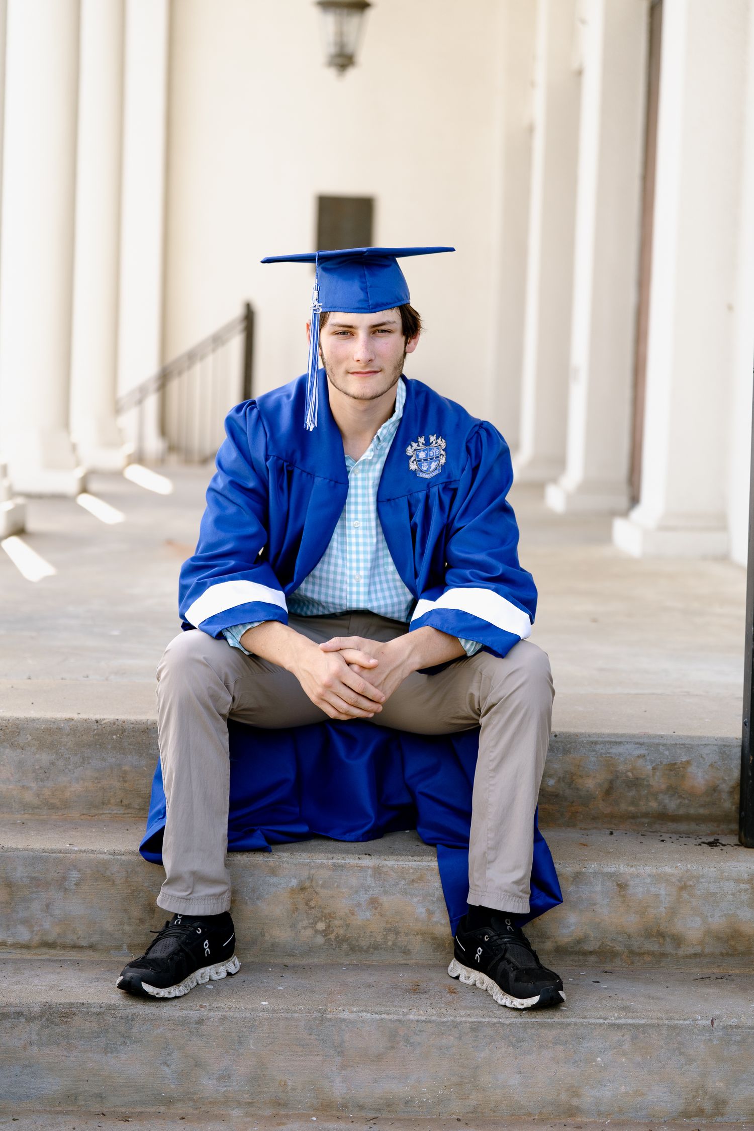 Graduate in blue cap and gown sits casually on steps with white columns in background.