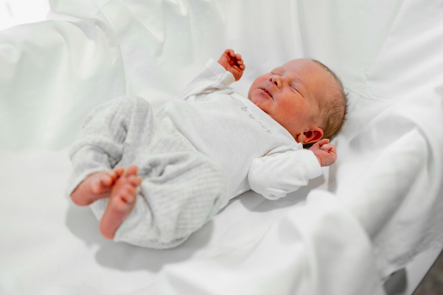 Tiny newborn in white sleepwear stretched out on white background.