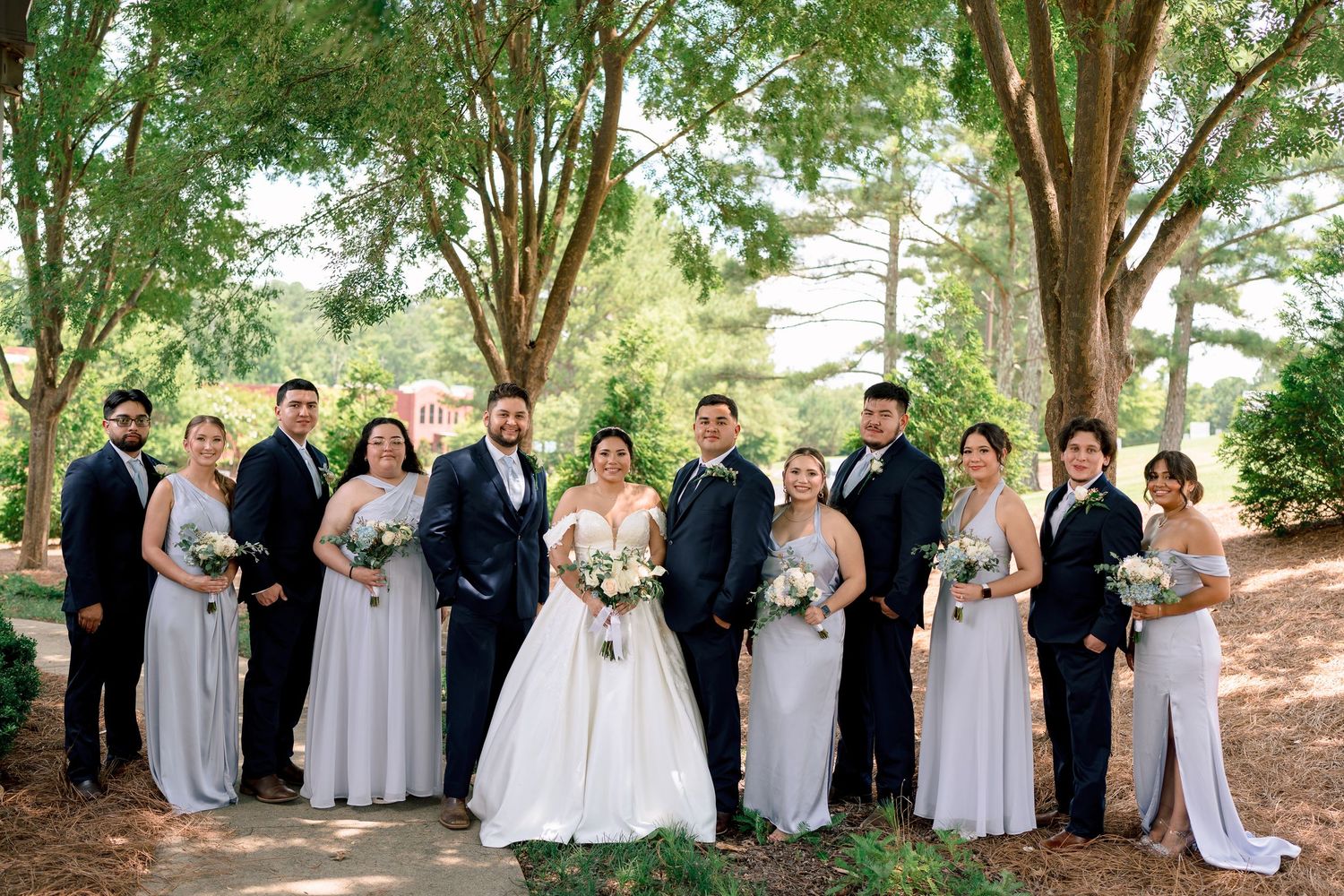 A wedding party in formal attire poses together under a canopy of trees in an outdoor setting.