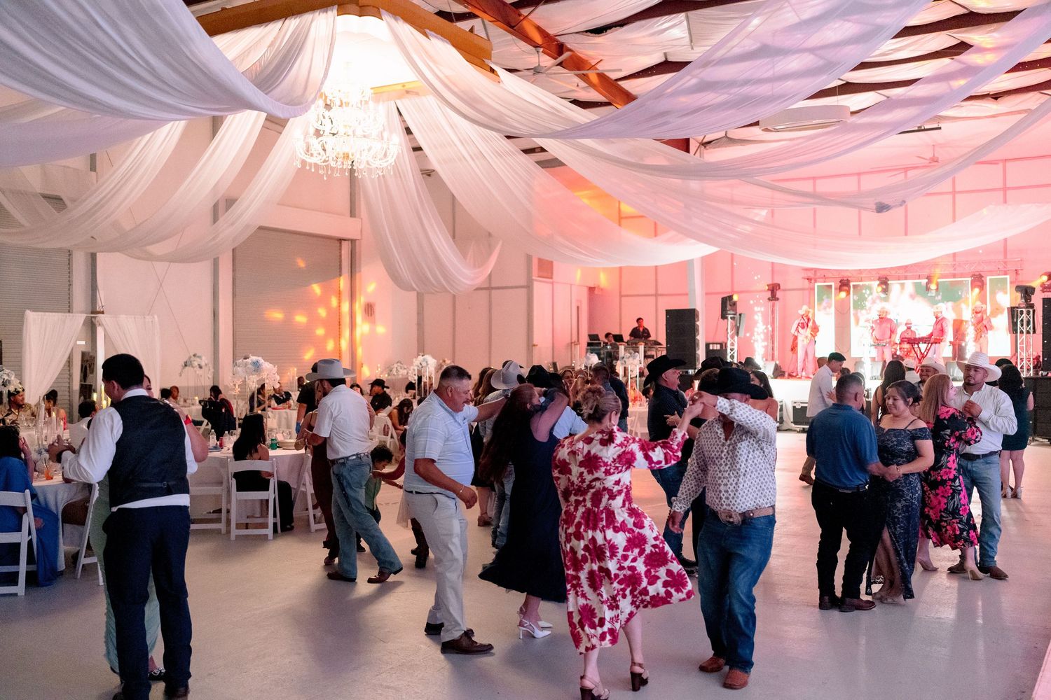 Wedding reception guests dance and socialize in an elegant white tent decorated with pink lighting and fabric draping.