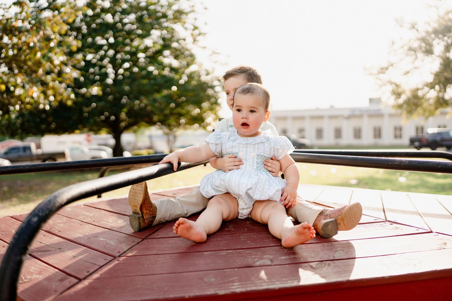 Two small children sit together on a red surface during an outdoor photo session.