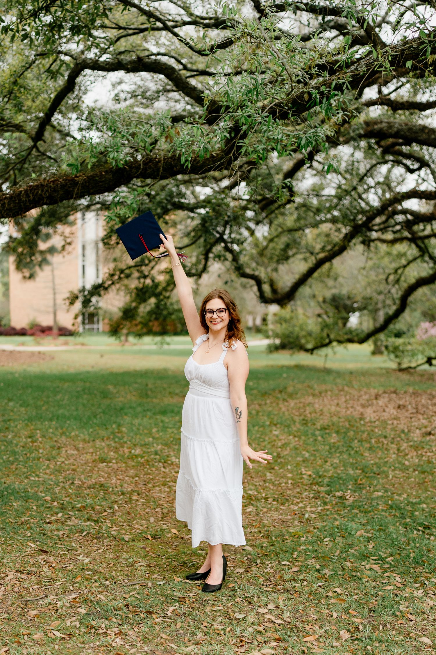 Graduate in a white dress poses under a sprawling oak tree during a graduation photoshoot.