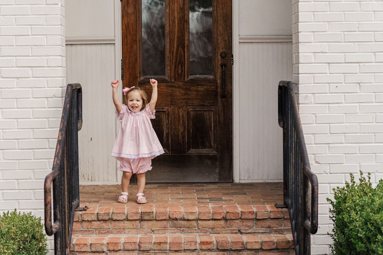 Joyful child in pink dress raises arms while standing on brick steps.