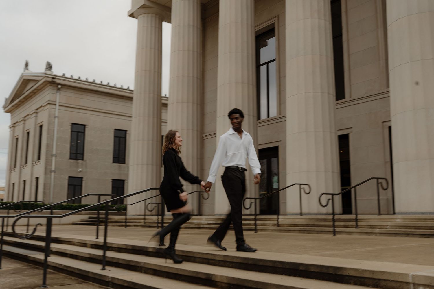 Couple holding hands walking down steps of a grand columned building in formal attire.