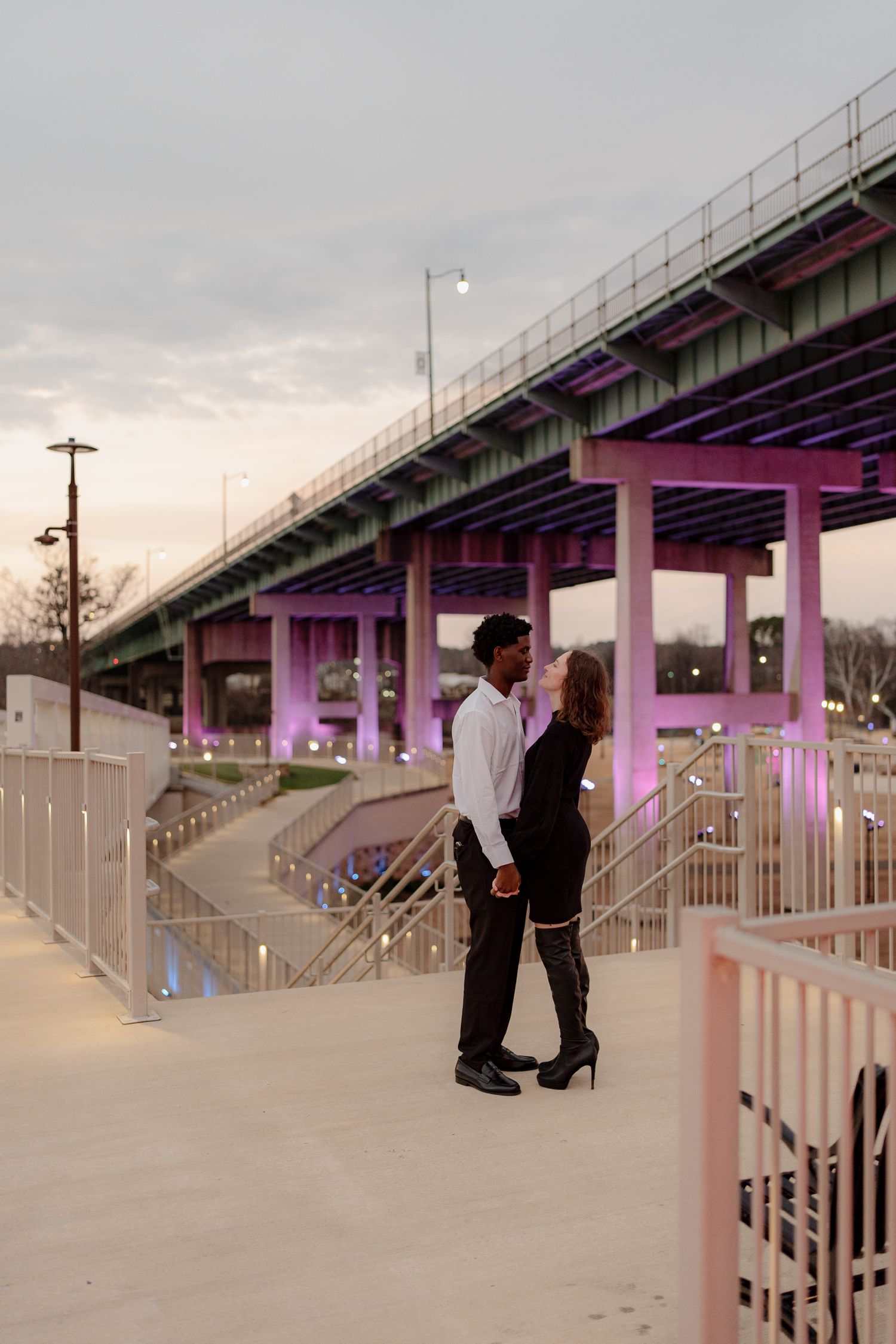 Couple standing close together on a bridge walkway illuminated by purple lights at dusk.
