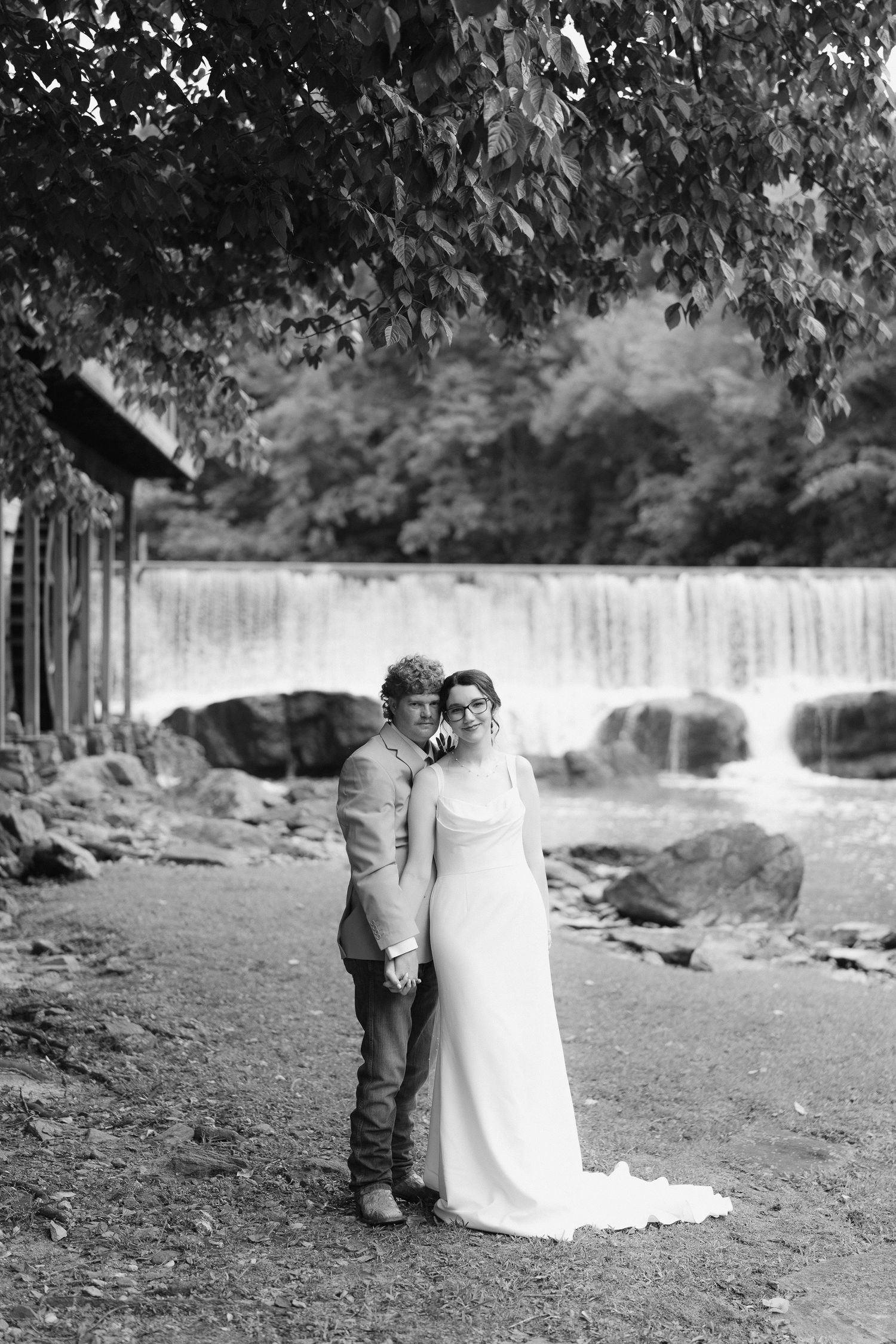 Black and white photo of bride and groom embracing near a waterfall and rocky riverbank.