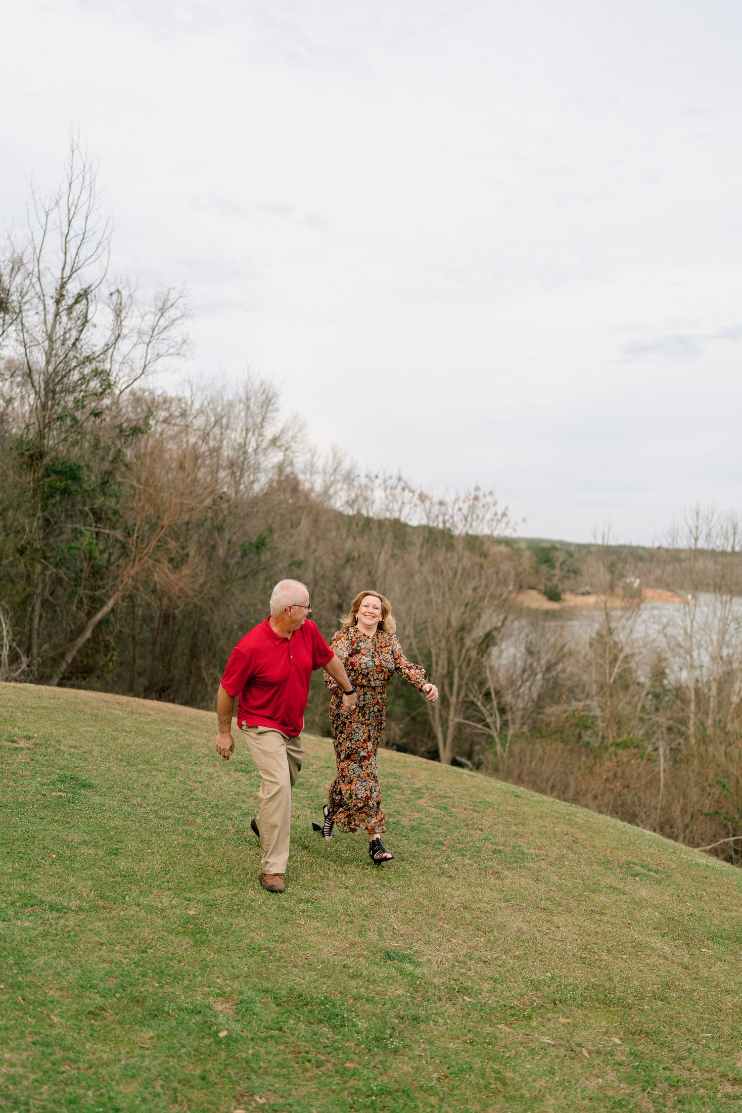Happy couple running and holding hands on a grassy hill with a lake and trees in background.