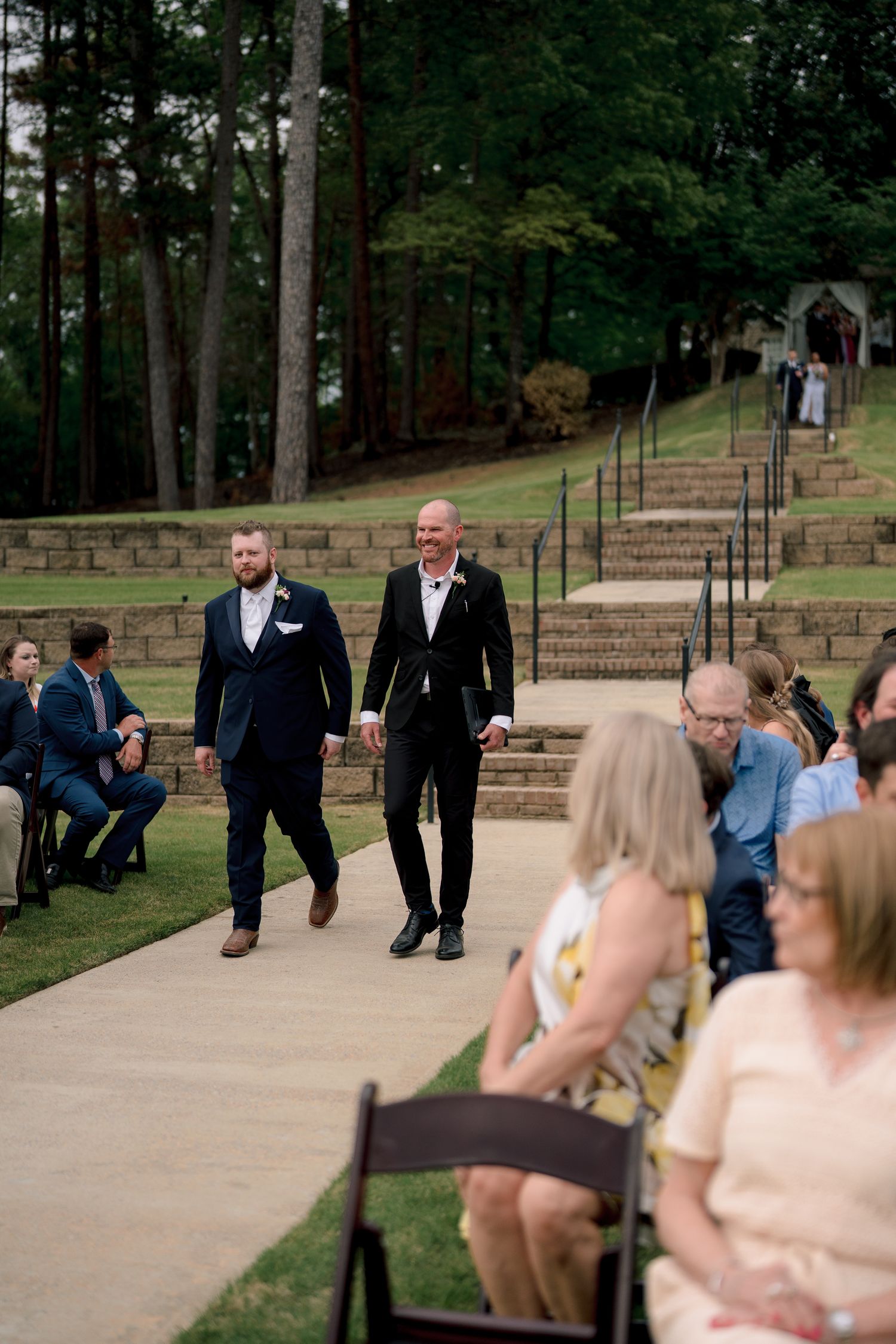 Two grooms walk hand-in-hand down the aisle at an outdoor wedding ceremony surrounded by guests.