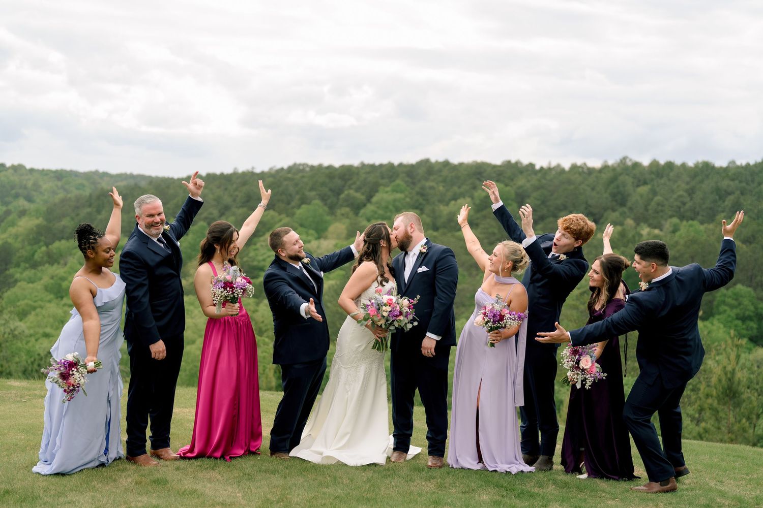 Joyful wedding party celebrates outdoors raising hands with green forest scenery in background.