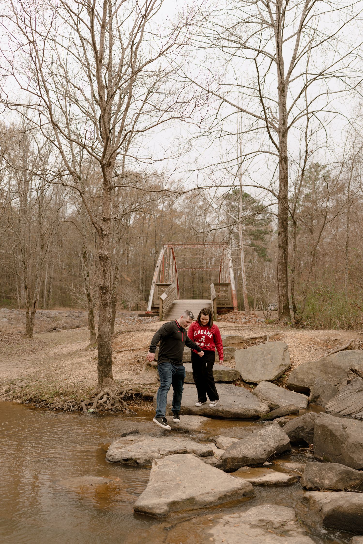 Two people sit on rocky outcroppings near a stream in a bare winter forest.