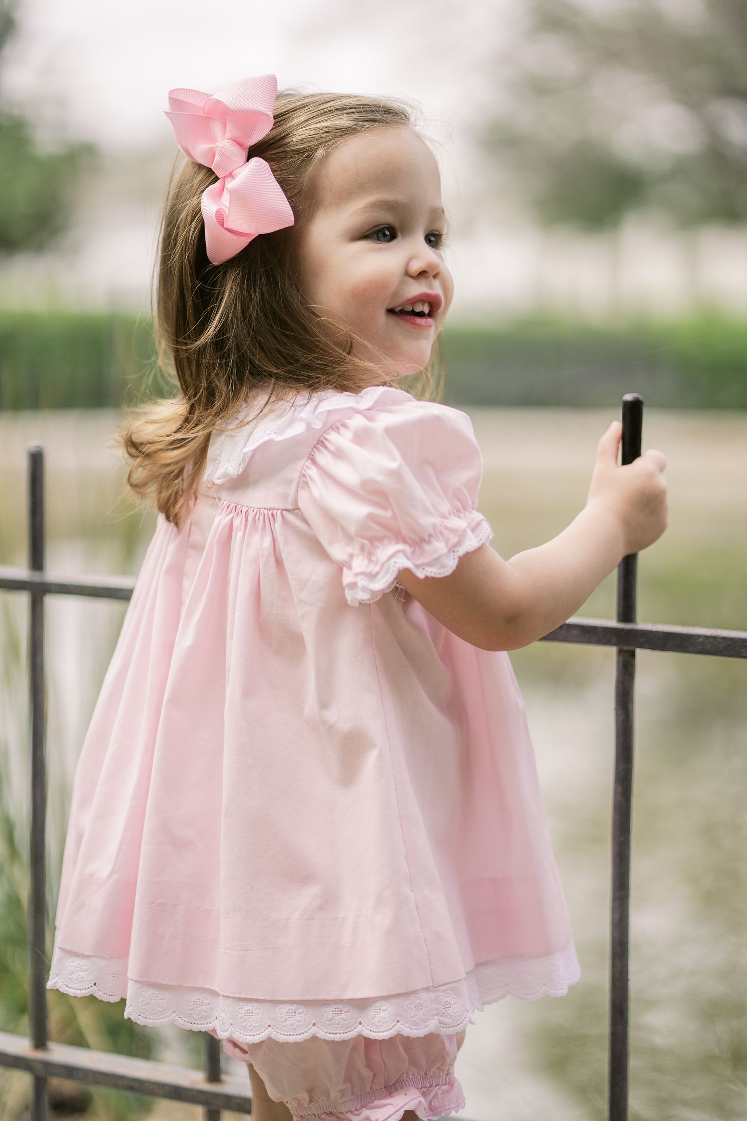 Child in light pink dress with ruffles and bow accessory poses while holding onto metal fence.
