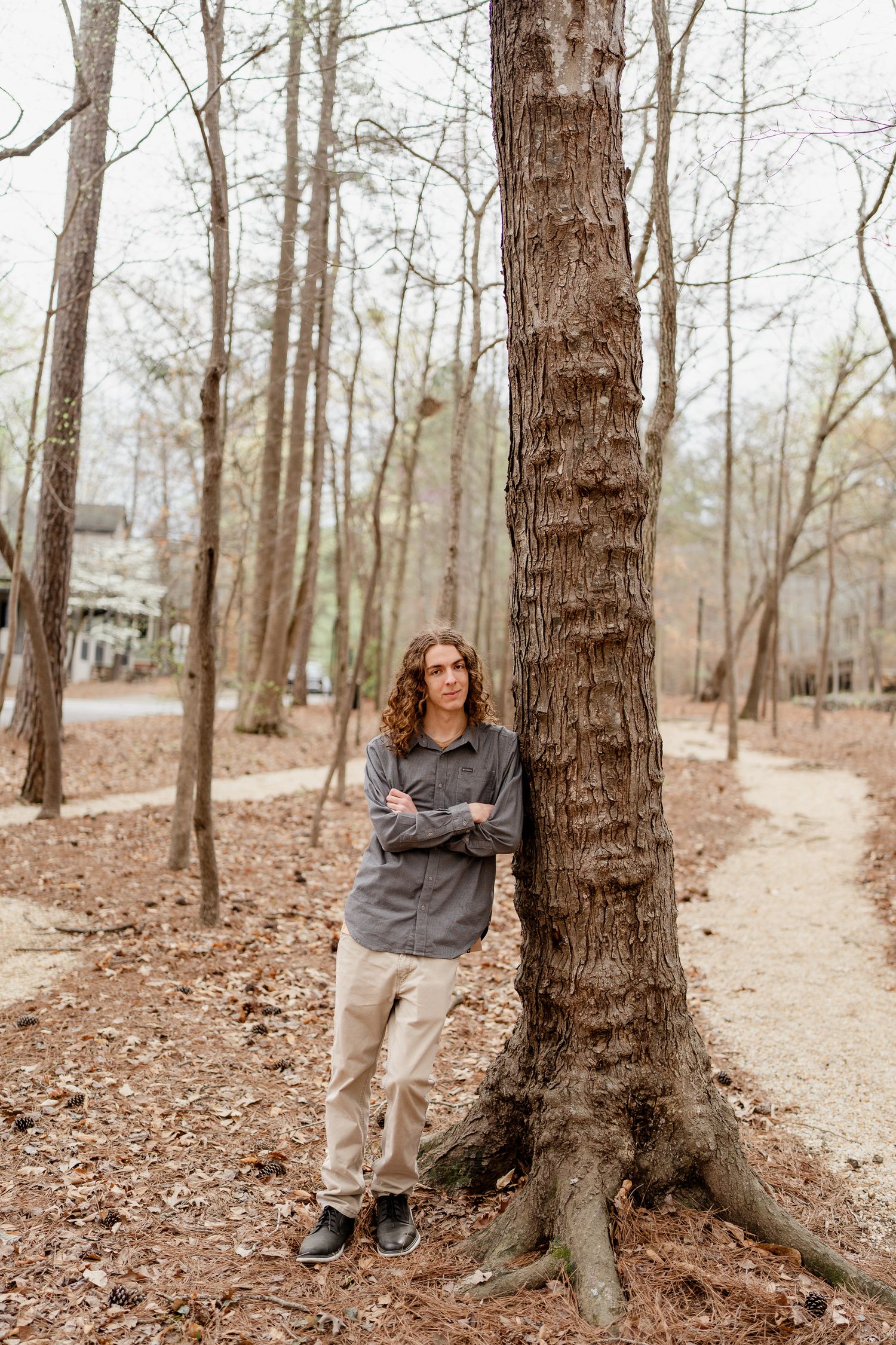 Person in casual attire leaning against large tree trunks in a wooded setting.