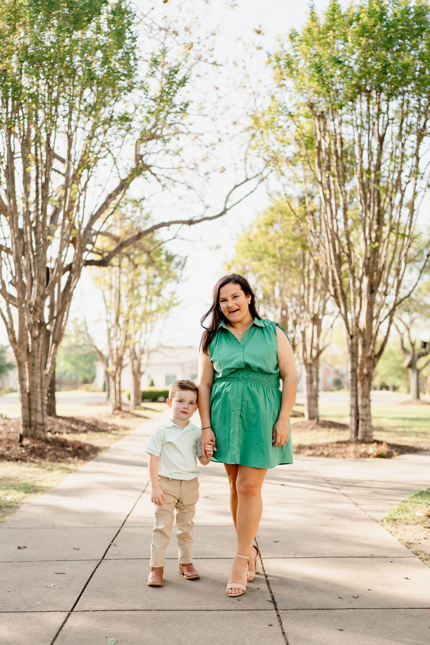 A woman in a green dress walks hand in hand with a child along a tree-lined path.