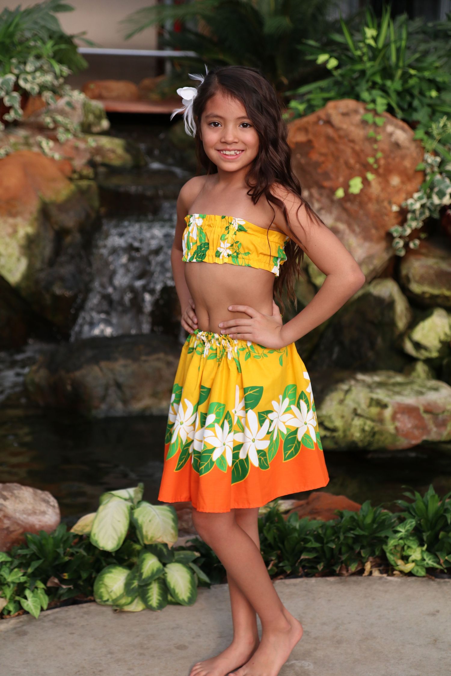 Child wearing a colorful yellow and orange tropical dress twirls in an outdoor garden setting.