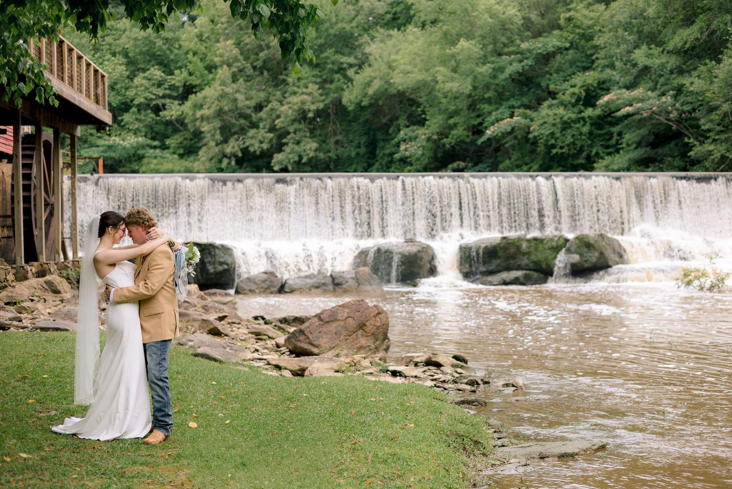Wedding couple embracing by a scenic waterfall and river surrounded by lush greenery.