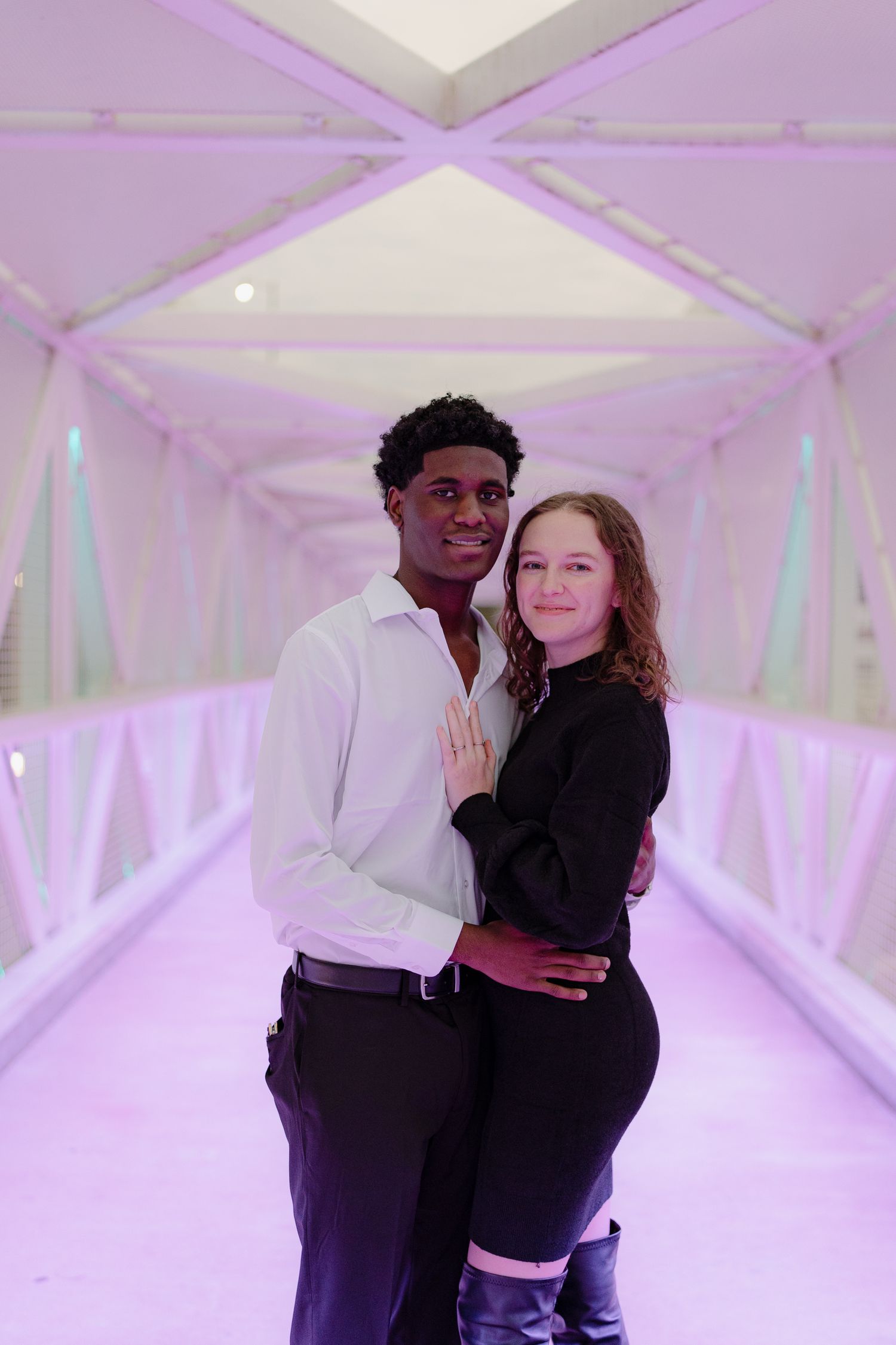 An interracial couple poses together on a purple-lit enclosed bridge, smiling at the camera.