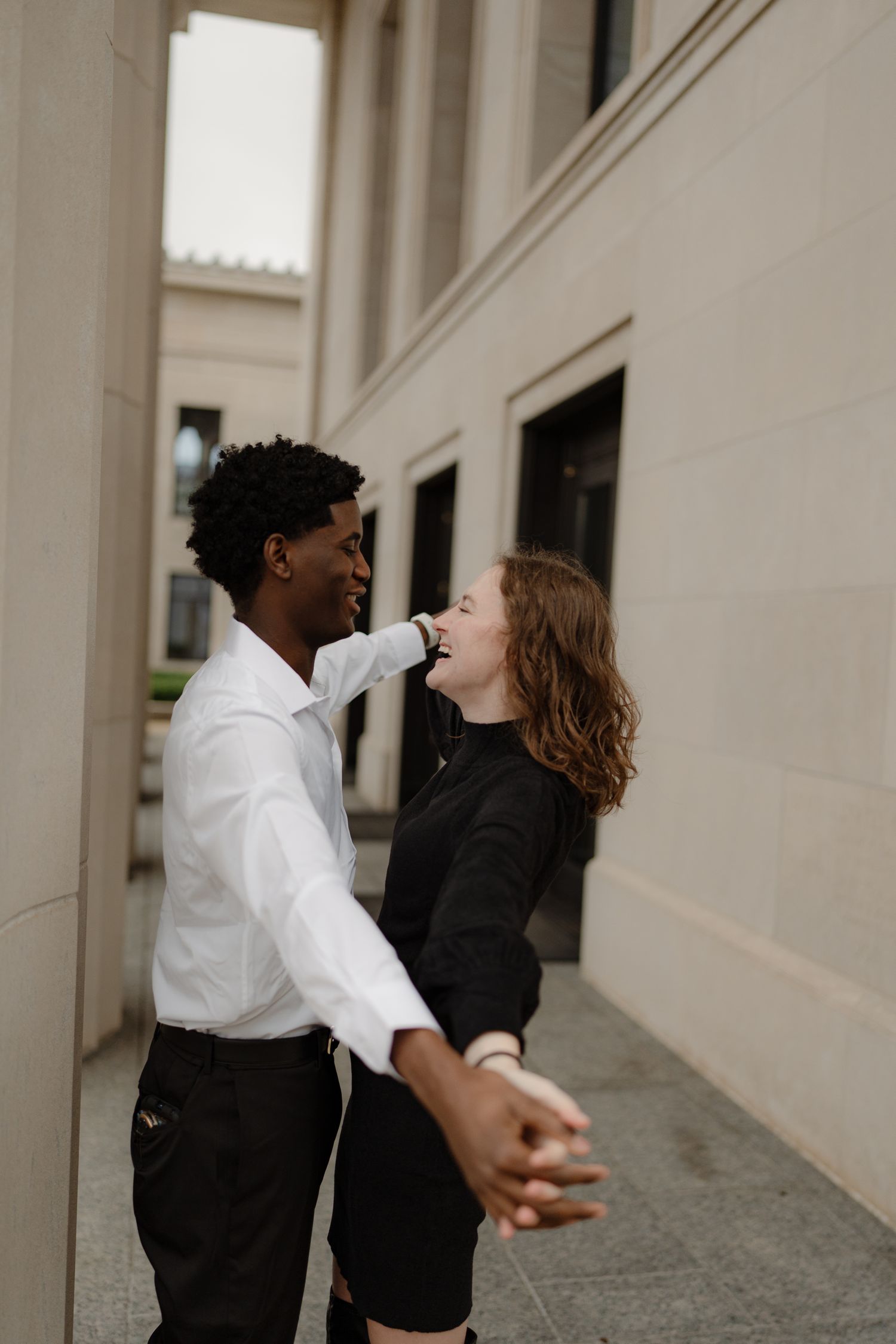 Joyful interracial couple dancing and laughing together outside a classic building, man in white shirt and woman in black dress.