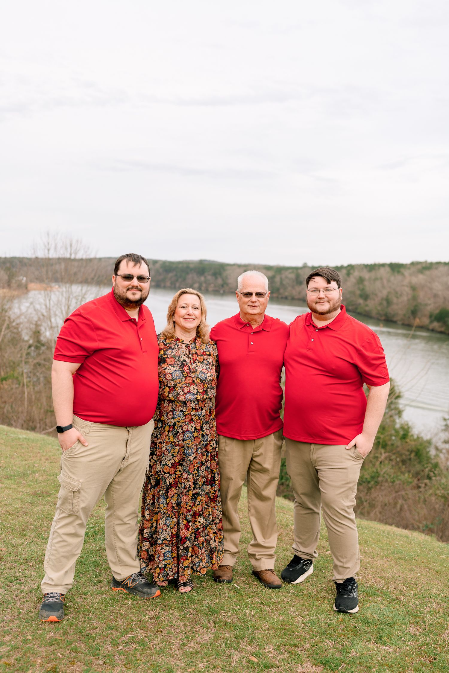 Family of four posing outdoors in matching red shirts and khakis with a scenic river and trees in the background.