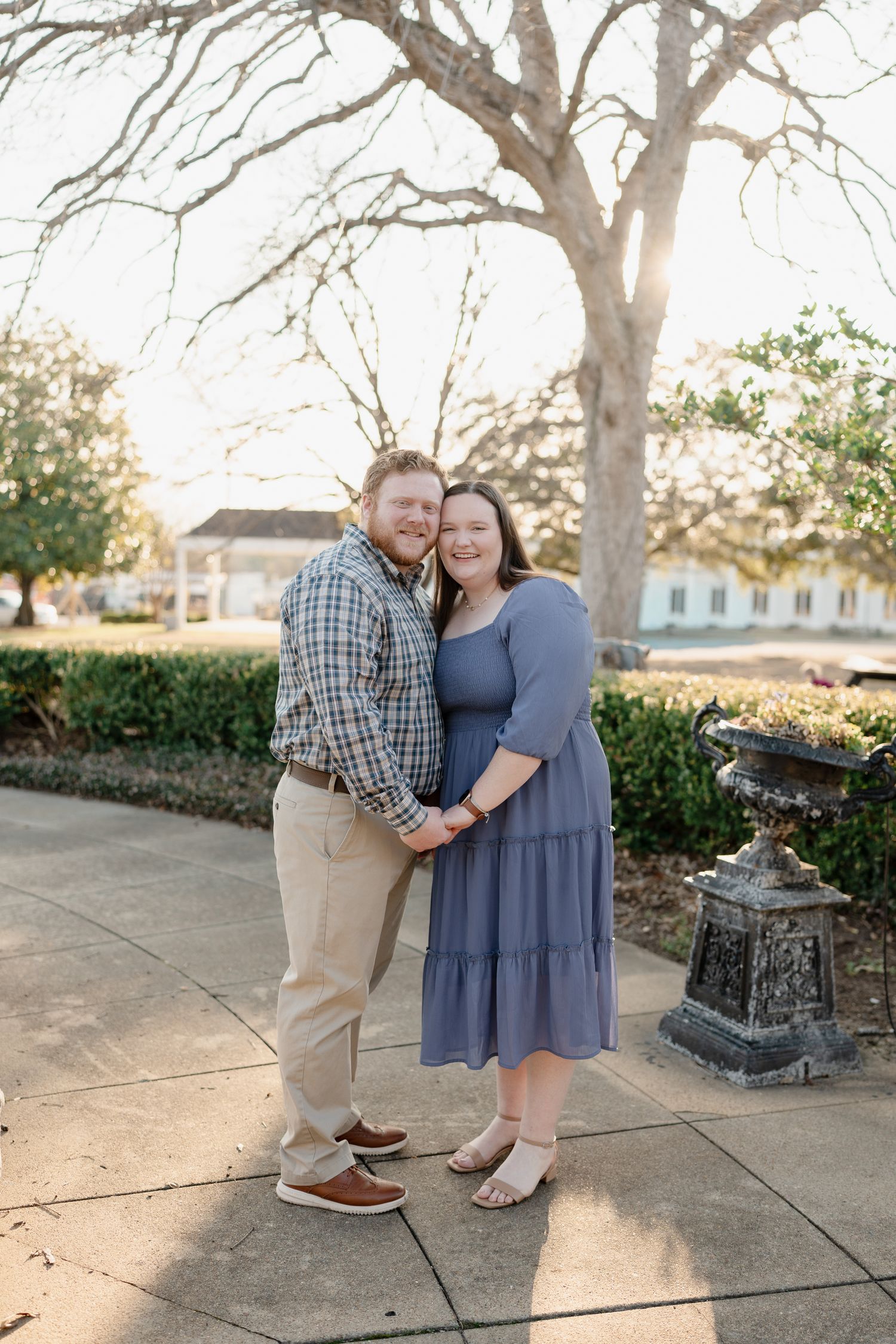 Couple posing together in a garden, woman in blue dress and man in plaid shirt, with trees and white building behind.