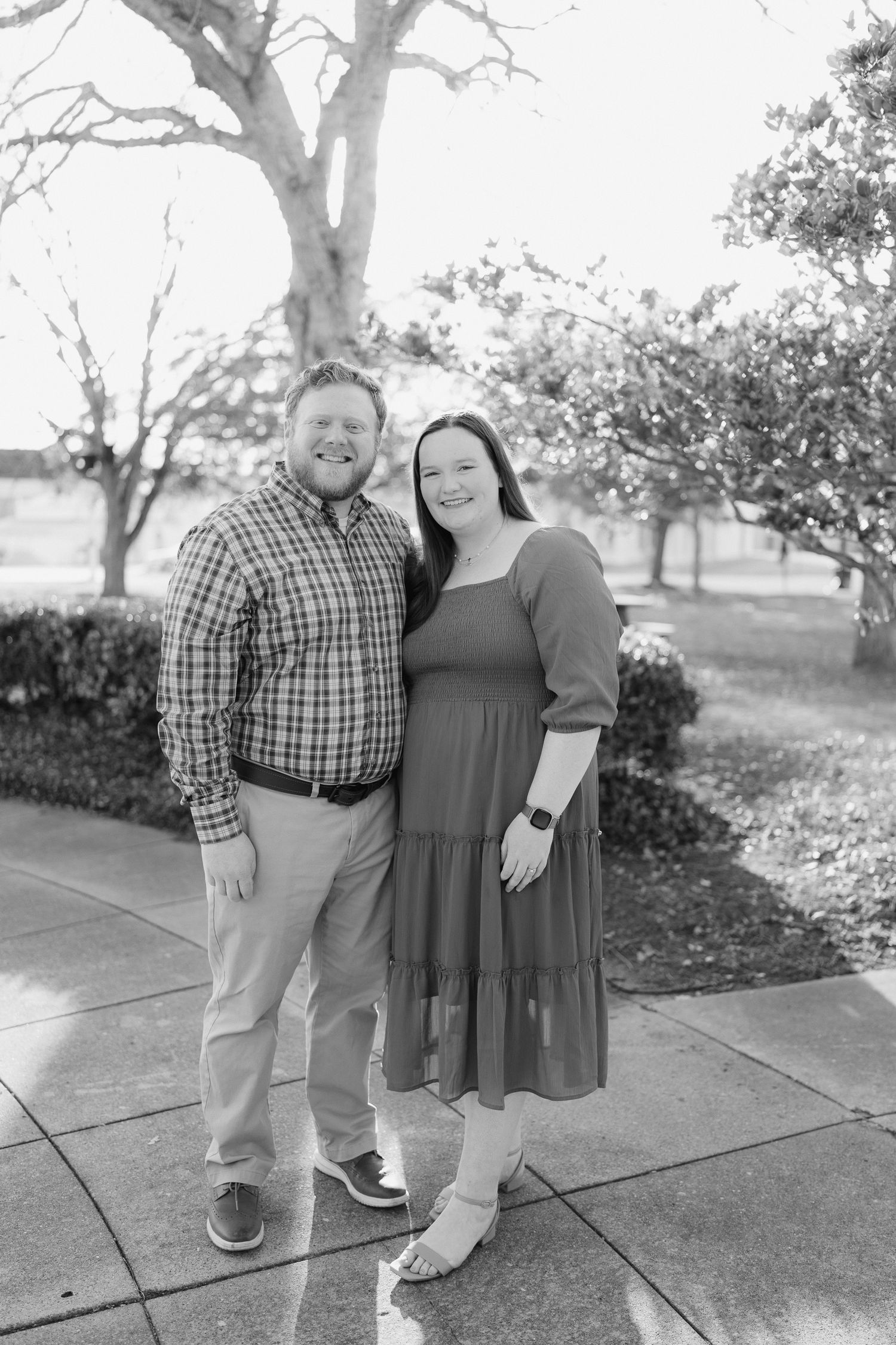 Smiling couple posing together on a sidewalk surrounded by trees and shrubs in black and white.